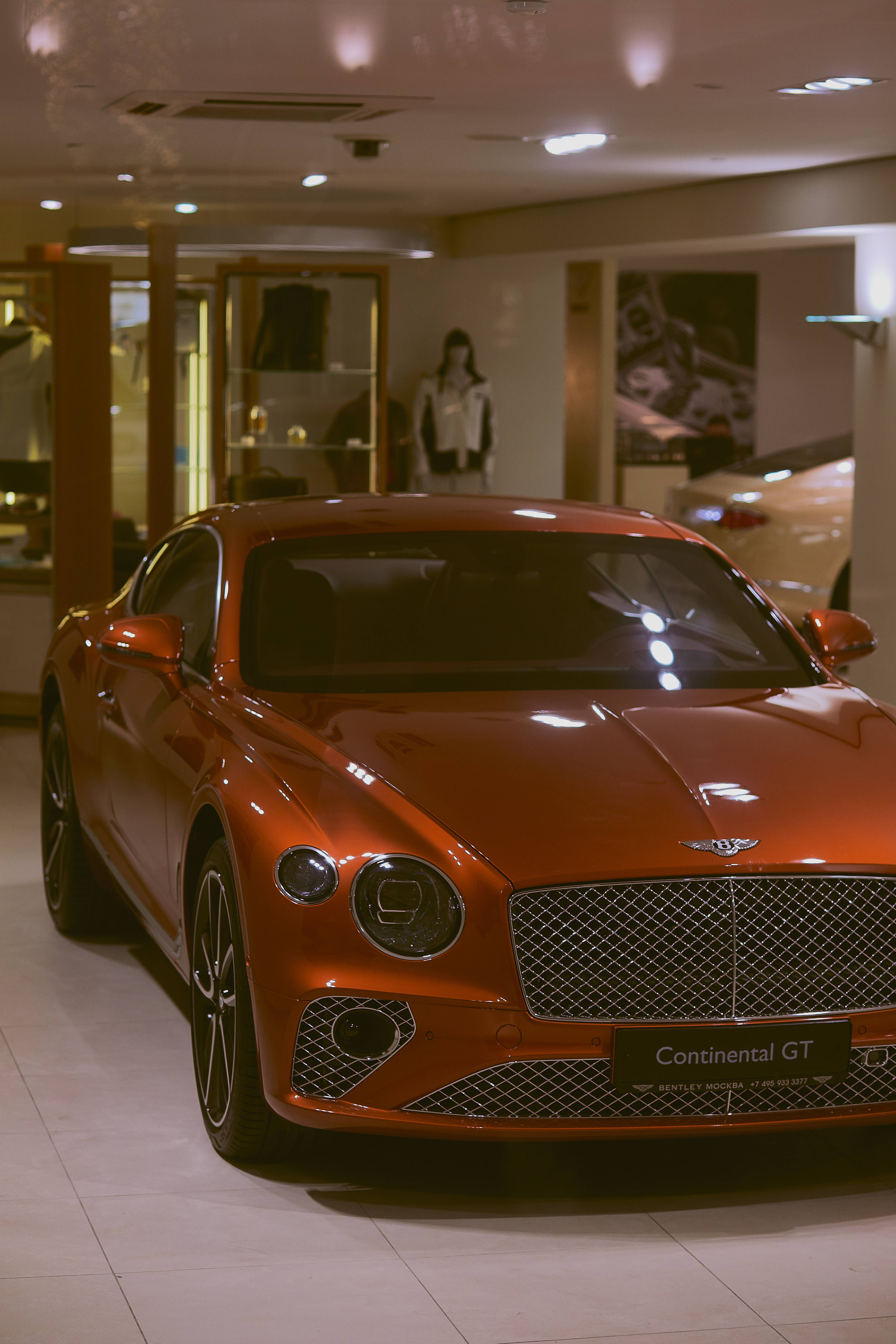 賓利 桌布 壁紙 — 一輛亮橙色的賓利 Continental GT 在展廳燈光下閃閃發光，反射光在彎曲的引擎蓋上舞動。
