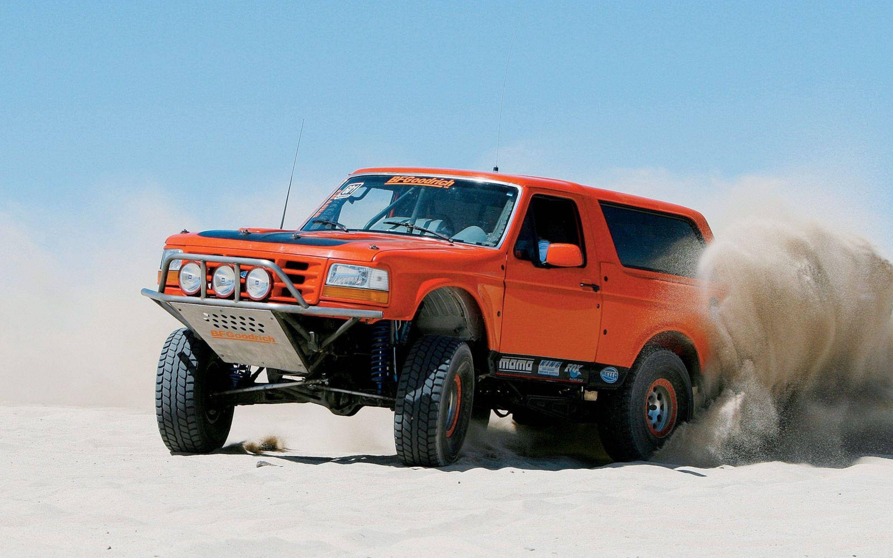 Ford Bronco 桌布 壁紙 — 一輛亮橙色的越野卡車在晴朗的藍天下，高速穿越廣闊的沙漠，揚起沙塵。