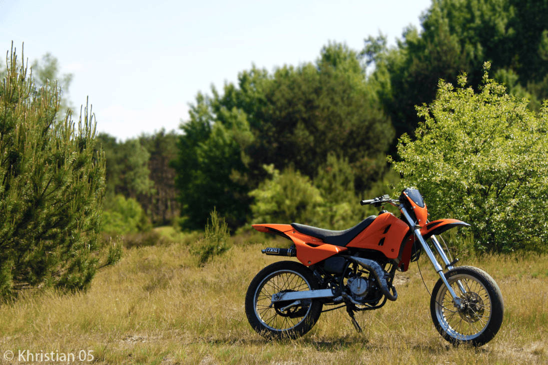 KTM 摩托車 桌布 壁紙 — 一輛亮橙色越野摩托車停在高草叢中，旁邊有綠樹，天空晴朗。