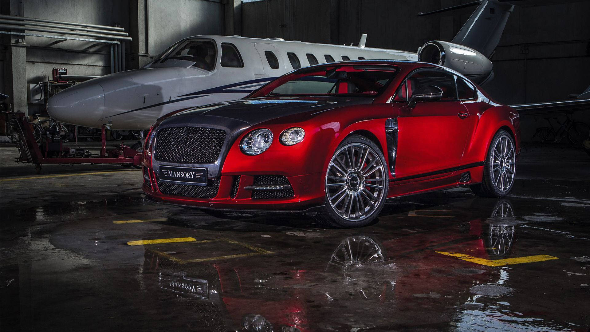 飛機 桌布 壁紙 — 一輛亮紅色 Mansory Bentley Continental GT 停放在潮濕的機庫中，旁邊有一架私人飛機，地板上有倒影。