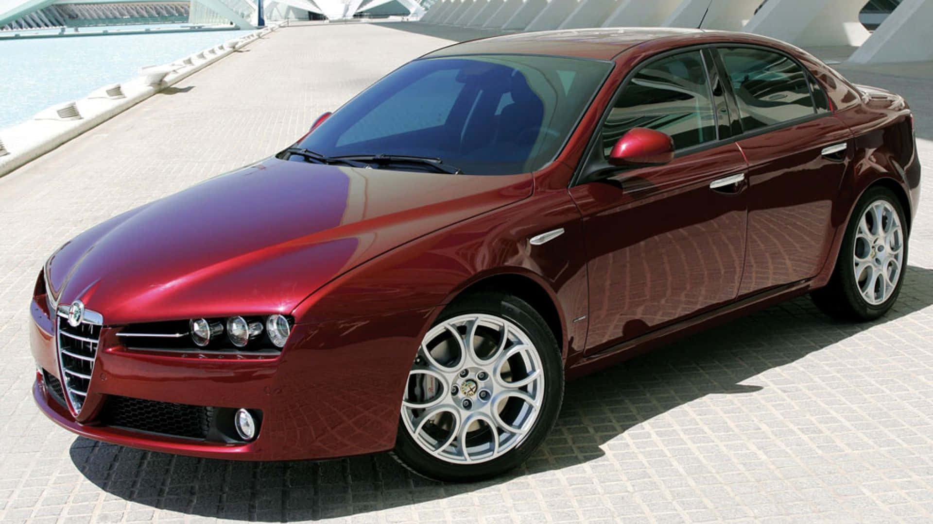 Alfa Romeo 159 桌布 壁紙 — 一輛亮紅色Alfa Romeo轎車停放在磁磚廣場上，旁邊是現代建築和水景。