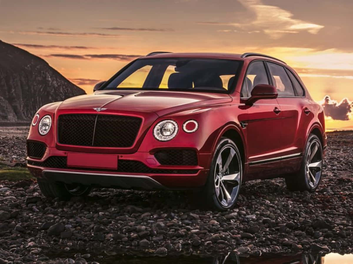 賓利 桌布 壁紙 — 一輛亮紅色Bentley Bentayga SUV停泊在岩石海灘上，背景是日落時分的山脈。