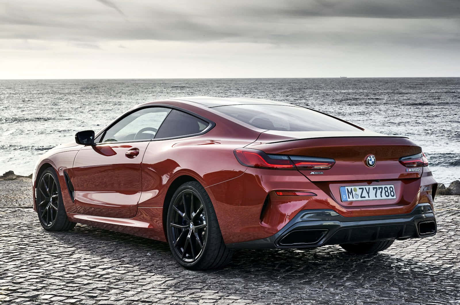 Bmw 8 Series 桌布 壁紙 — 一輛亮紅色BMW 8系列轎跑車停泊在鵝卵石海岸上，背後是廣闊的海洋和陰天。
