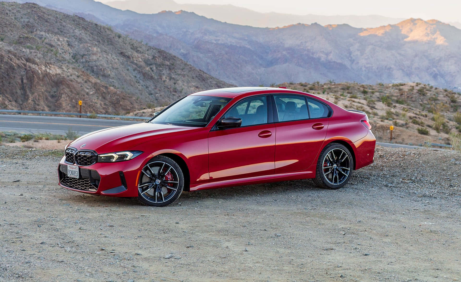 Bmw 3 Series 桌布 壁紙 — 一輛亮紅色BMW M3轎車停在沙漠公路旁的礫石上，崎嶇的山脈沐浴在金黃色的日落光芒中。