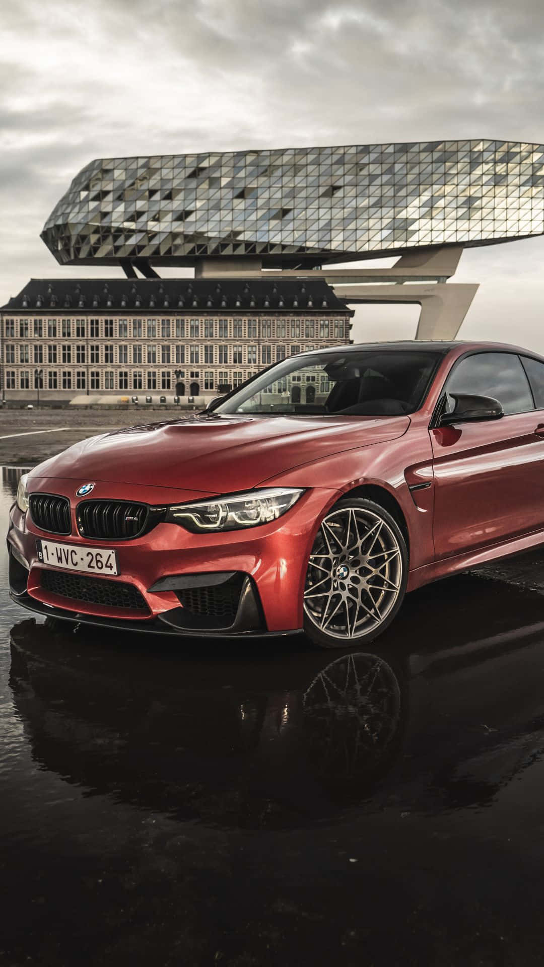 Android Bmw 桌布 壁紙 — 一輛亮紅色BMW M4轎跑車在潮濕路面上倒映，天空多雲，背後有一座現代幾何建築。