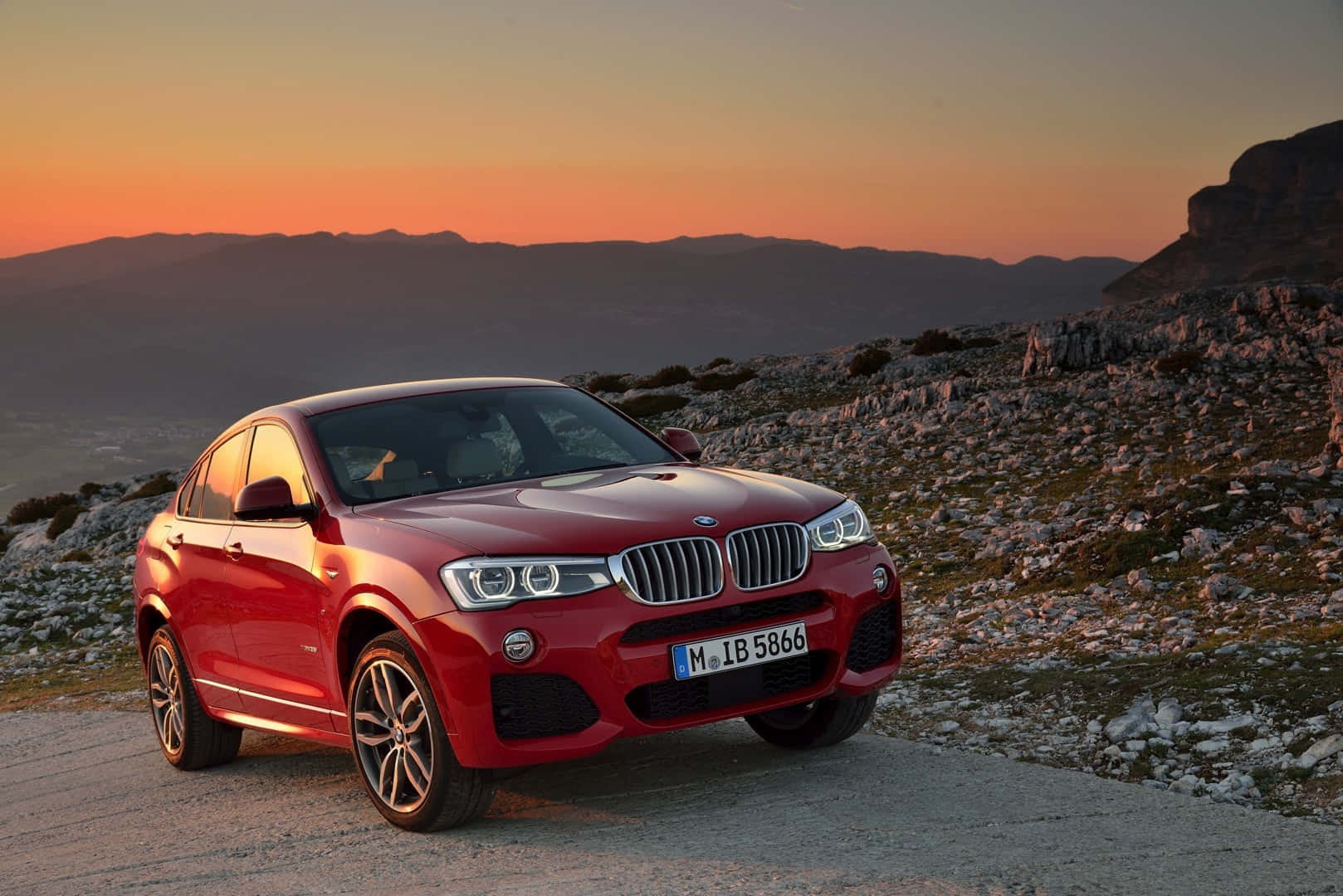 Bmw X4 桌布 壁紙 — 一輛亮紅色BMW X4 SUV停泊在日落時分的山路，天空橙黃，地形岩石。