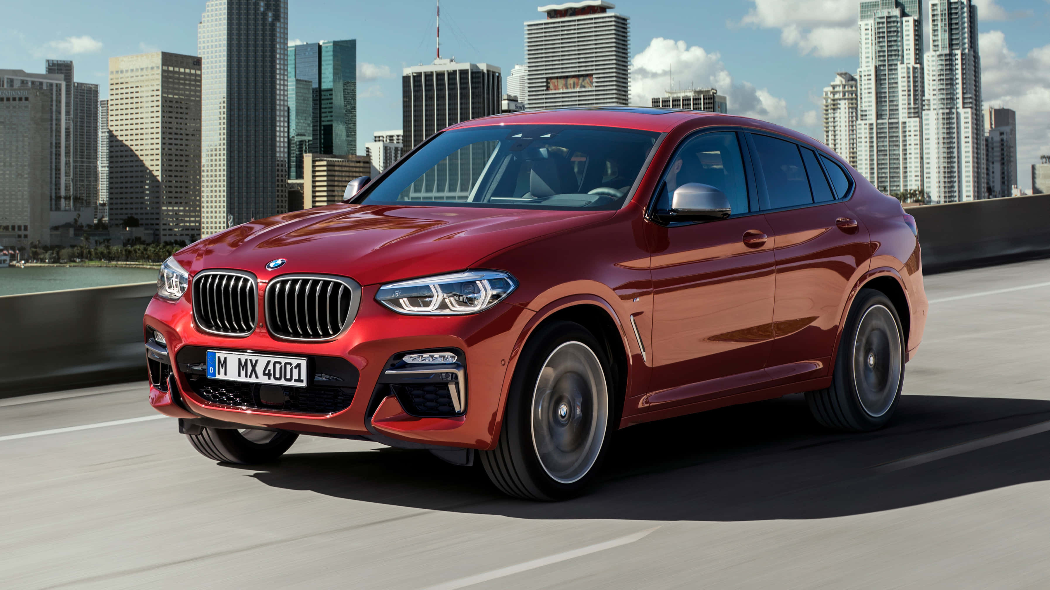 Bmw X4 桌布 壁紙 — 一輛亮紅色BMW X4在橋上行駛，背景是現代城市天際線和水面，天空湛藍明亮。