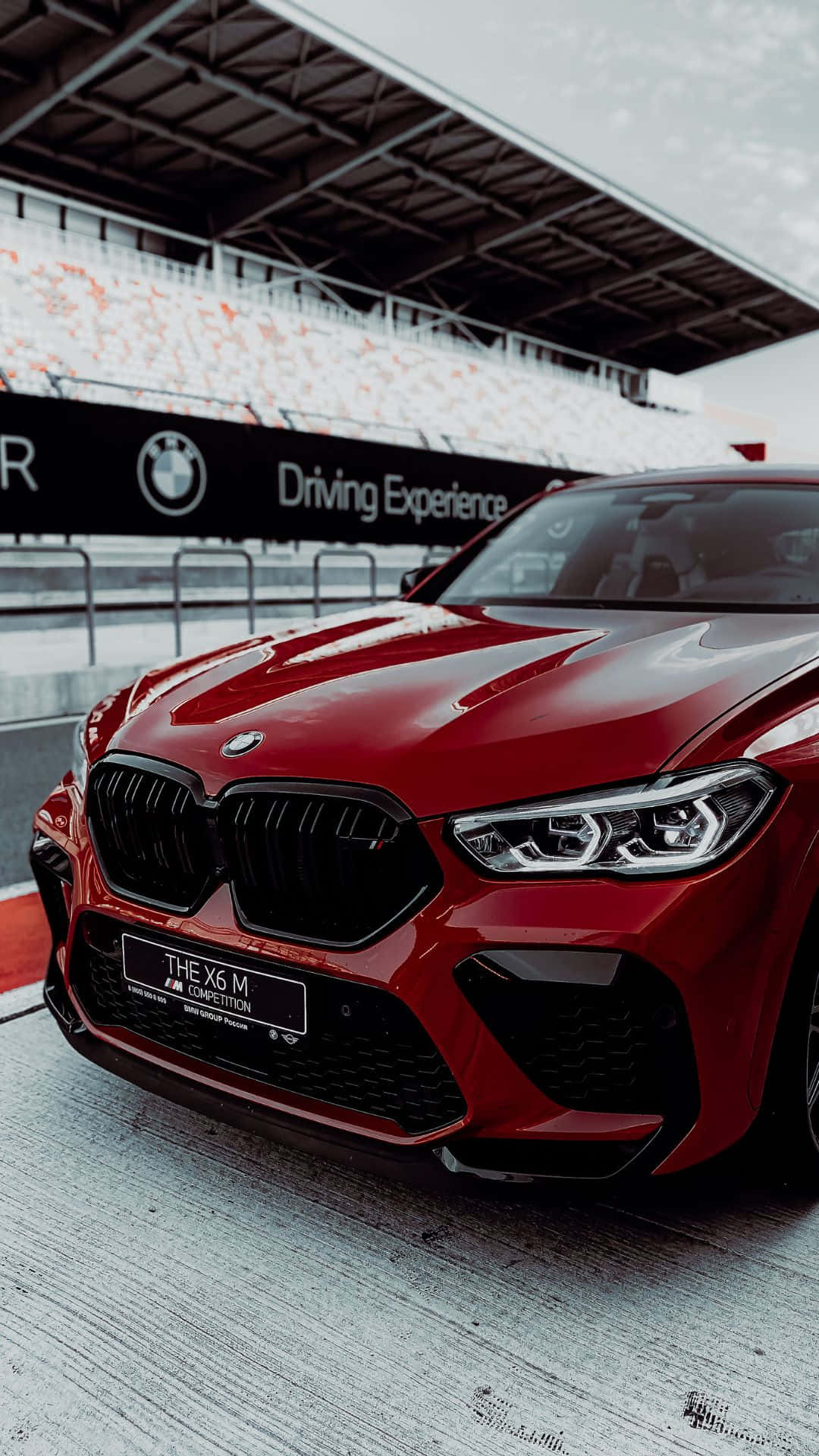 Android Bmw 桌布 壁紙 — 一輛亮紅色BMW X6 M Competition停泊在賽車場上，背景有看台，天空多雲。