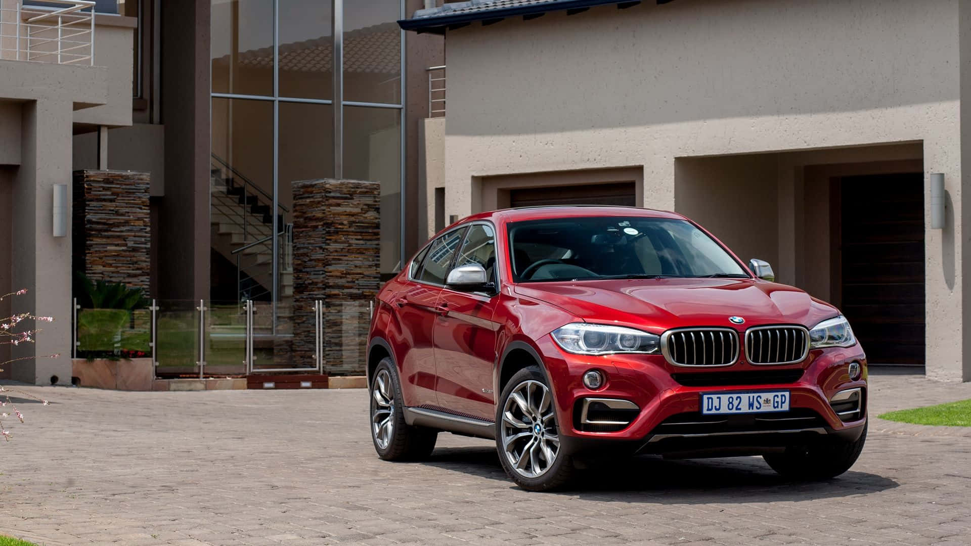 Bmw X6 桌布 壁紙 — 一輛亮紅色BMW X6 SUV停泊在現代化房屋前的石頭車道上，房屋有玻璃牆和石柱。