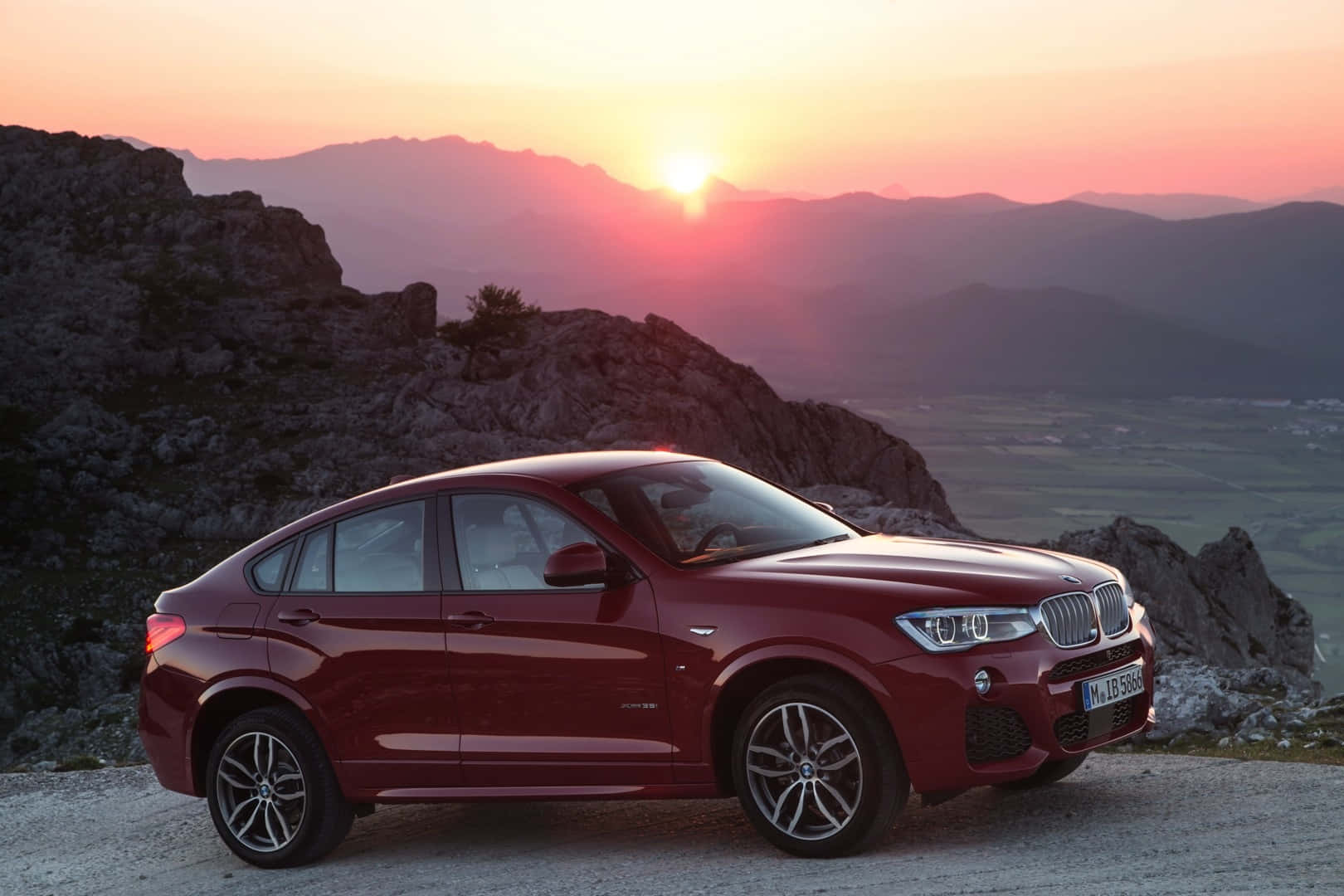 Bmw X4 桌布 壁紙 — 一輛亮紅色BMW X6停泊在岩石觀景點，夕陽在遠山後落下，天空染上溫暖的橙色和粉紅色調。