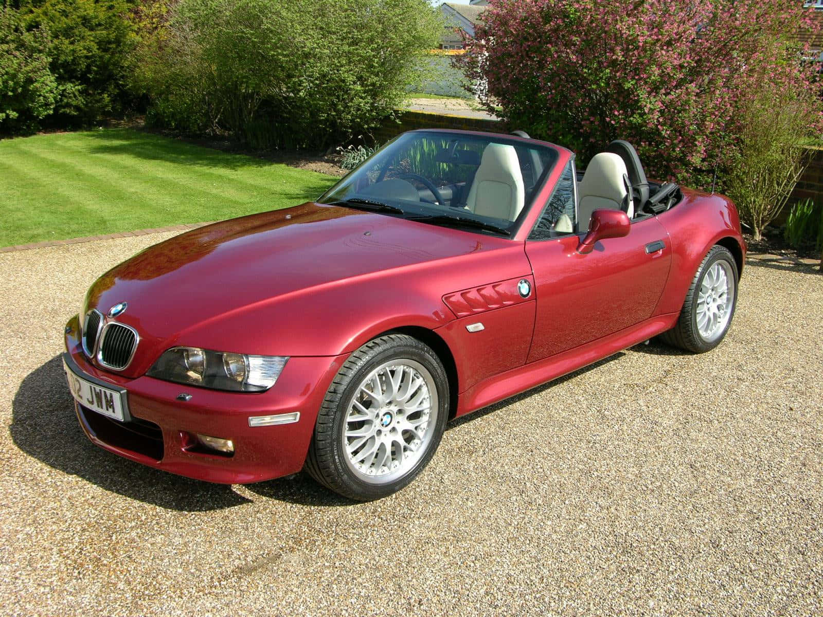 Bmw Z3 桌布 壁紙 — 一輛亮紅色BMW Z3敞篷跑車停在碎石路上，周圍有綠色草坪和開花的灌木叢，在明亮的日光下。