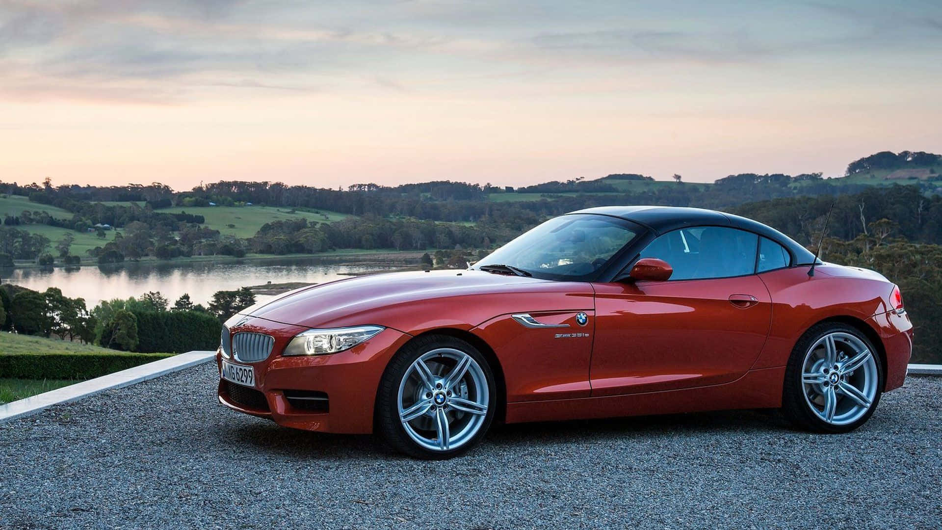 Bmw Z4 桌布 壁紙 — 一輛亮紅色BMW Z4敞篷跑車停泊在碎石上，俯瞰日落時分的寧靜湖泊，背景是起伏的綠色山丘。