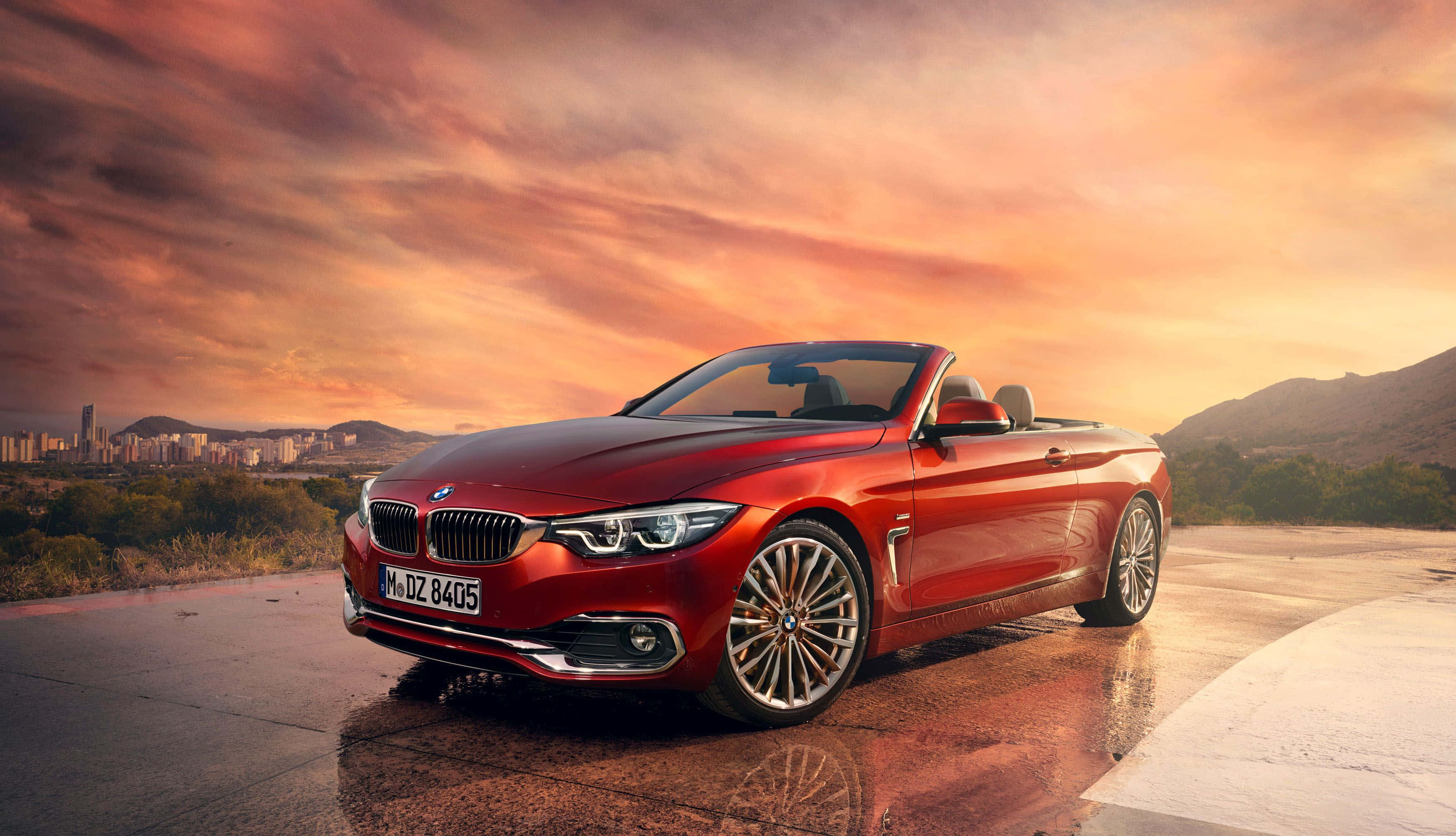 Bmw 4 Series 桌布 壁紙 — 一輛亮紅色BMW敞篷車停泊在日落時分的濕滑道路上，背景是城市景觀和山脈。