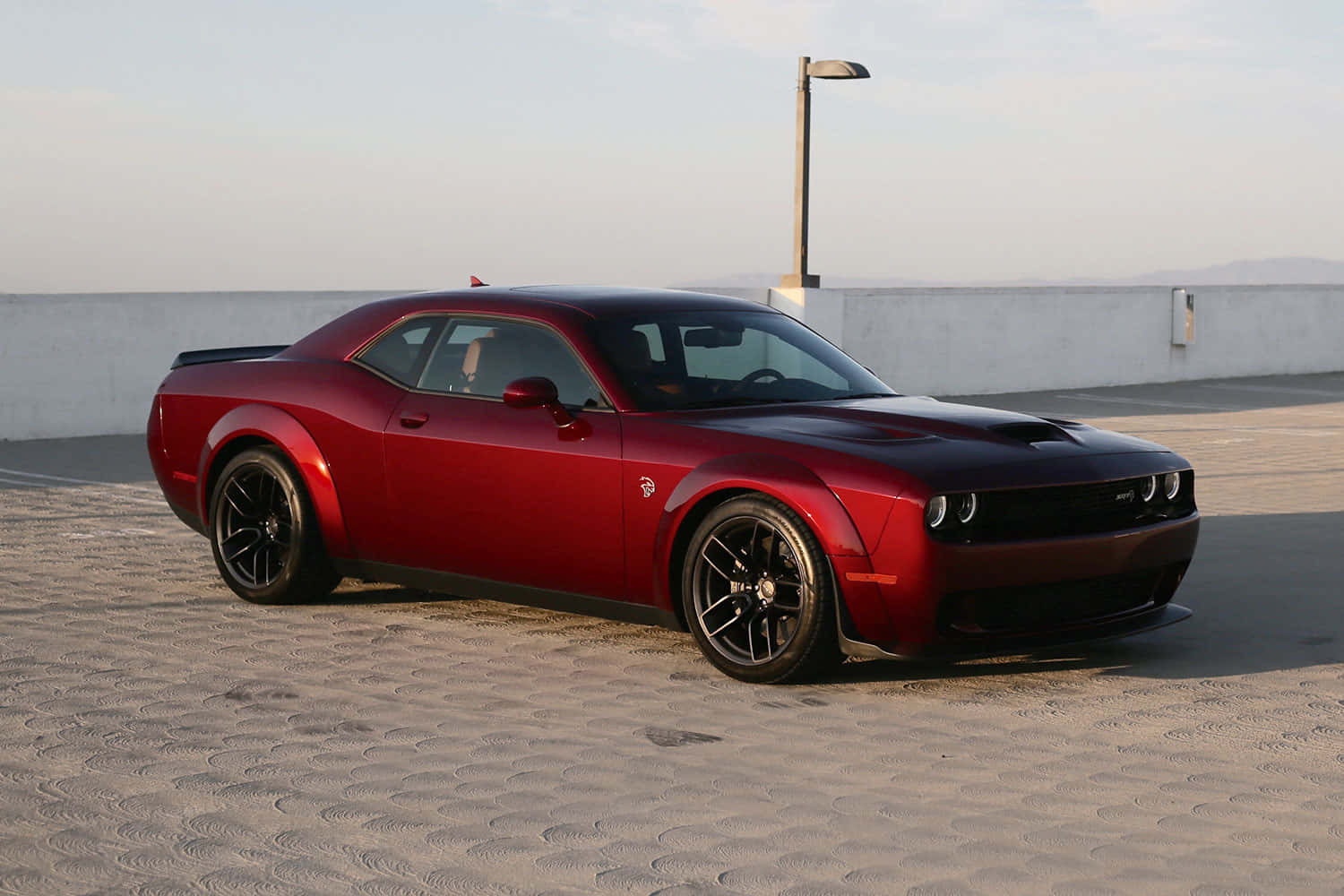 Dodge 桌布 壁紙 — 一輛亮紅色Dodge Challenger肌肉車停放在紋理路面上，柔和日光下，背景是簡約混凝土牆和遠山。