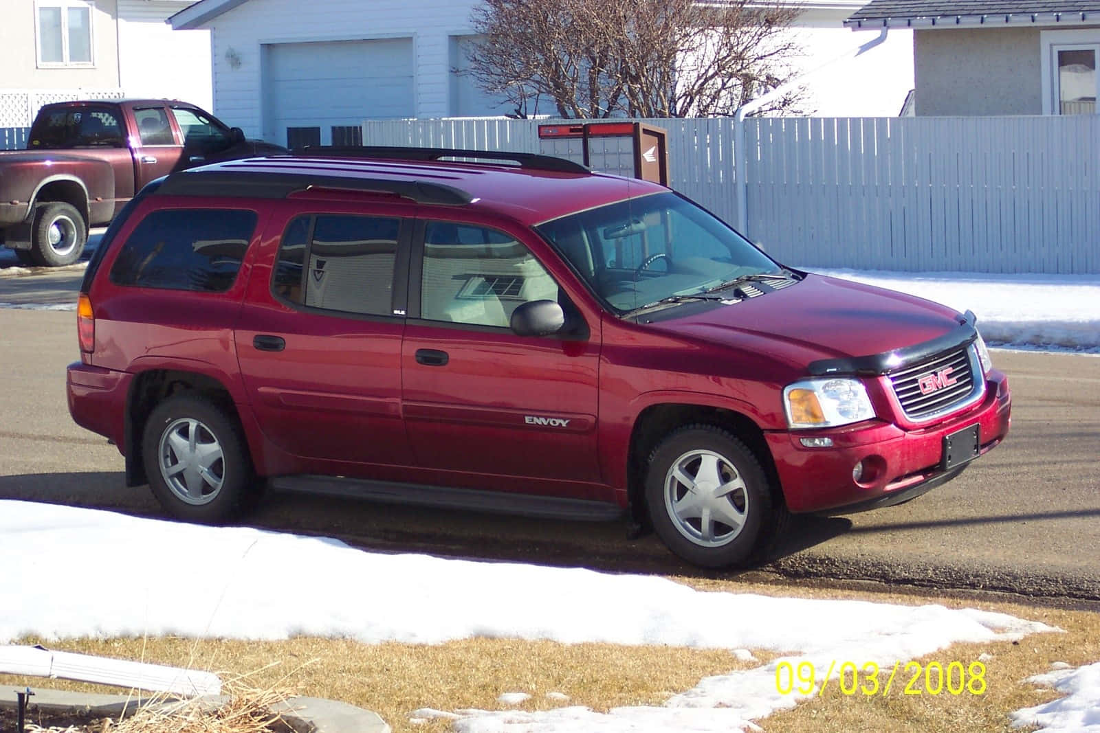 Gmc Envoy 桌布 壁紙 — 一輛亮紅色GMC Envoy SUV停放在積雪的住宅街道上，背景有白色圍欄和光禿禿的樹木。