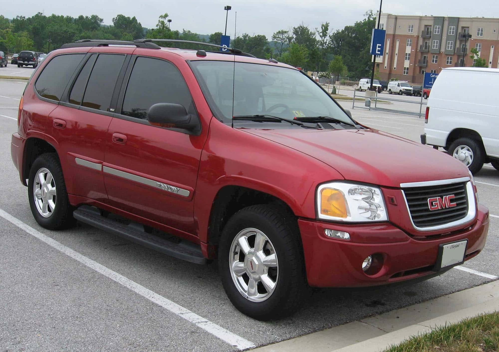 Gmc Envoy 桌布 壁紙 — 一輛亮紅色GMC Envoy SUV停放在鋪砌的停車場，背景有樹木和公寓大樓。