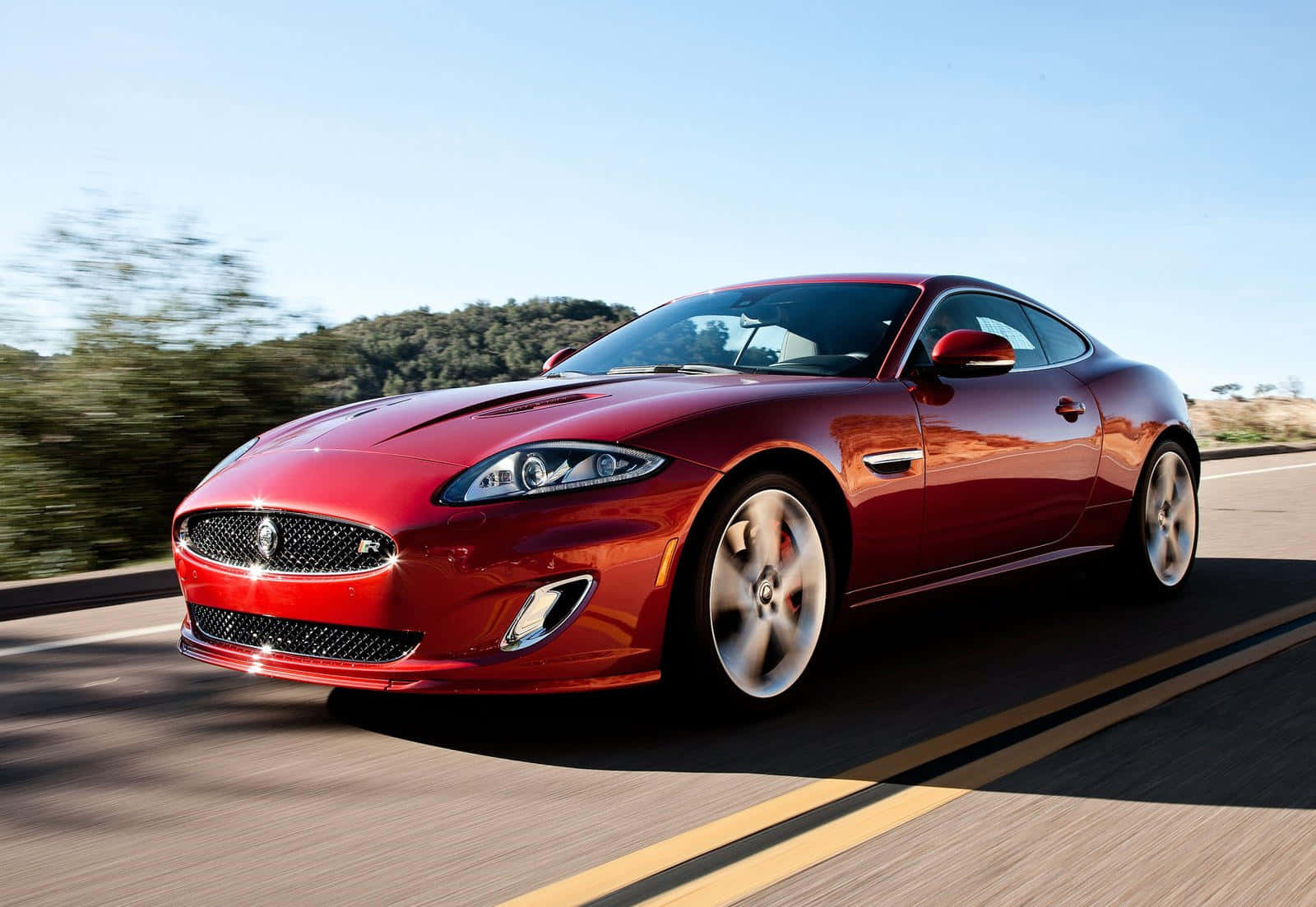 Jaguar Xkr 桌布 壁紙 — 一輛亮紅色Jaguar跑車在陽光明媚的高速公路上疾馳，背景是模糊的綠色山丘。