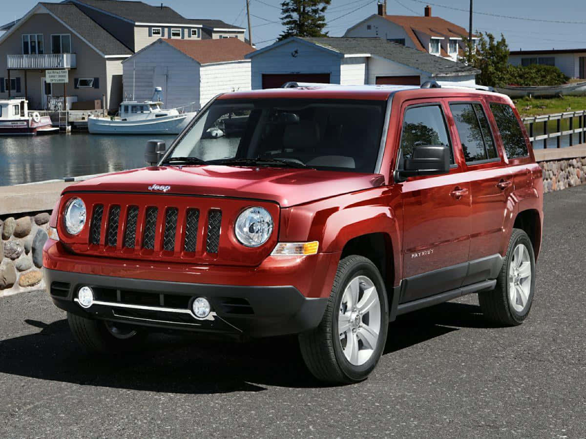 Jeep Patriot 桌布 壁紙 — 一輛亮紅色Jeep Patriot SUV停放在柏油路上，背景是陽光普照的海濱，有船隻和房屋。