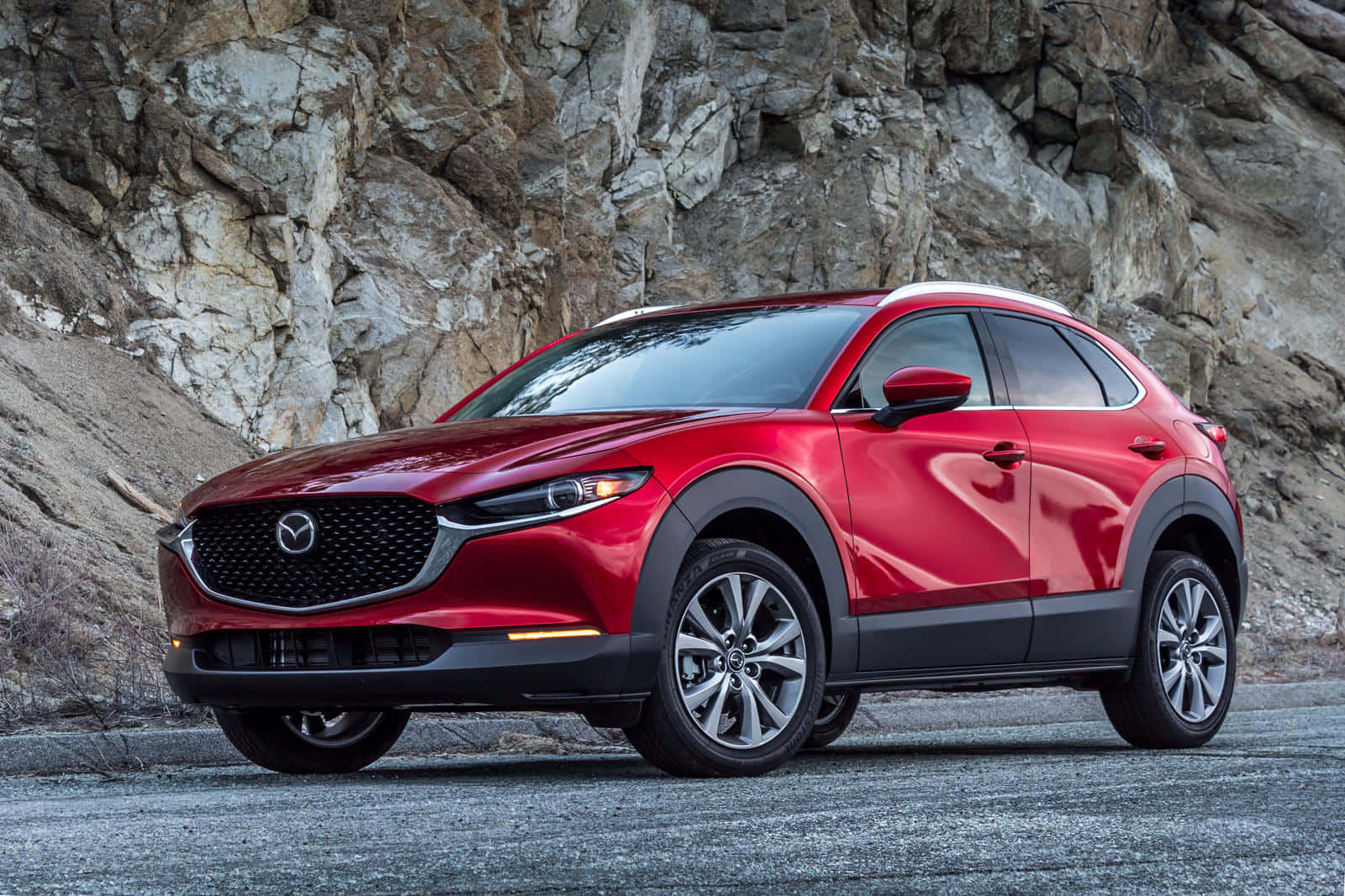 Mazda Cx-30 桌布 壁紙 — 一輛亮紅色Mazda CX-30 SUV停泊在崎嶇岩石懸崖前的碎石路上。