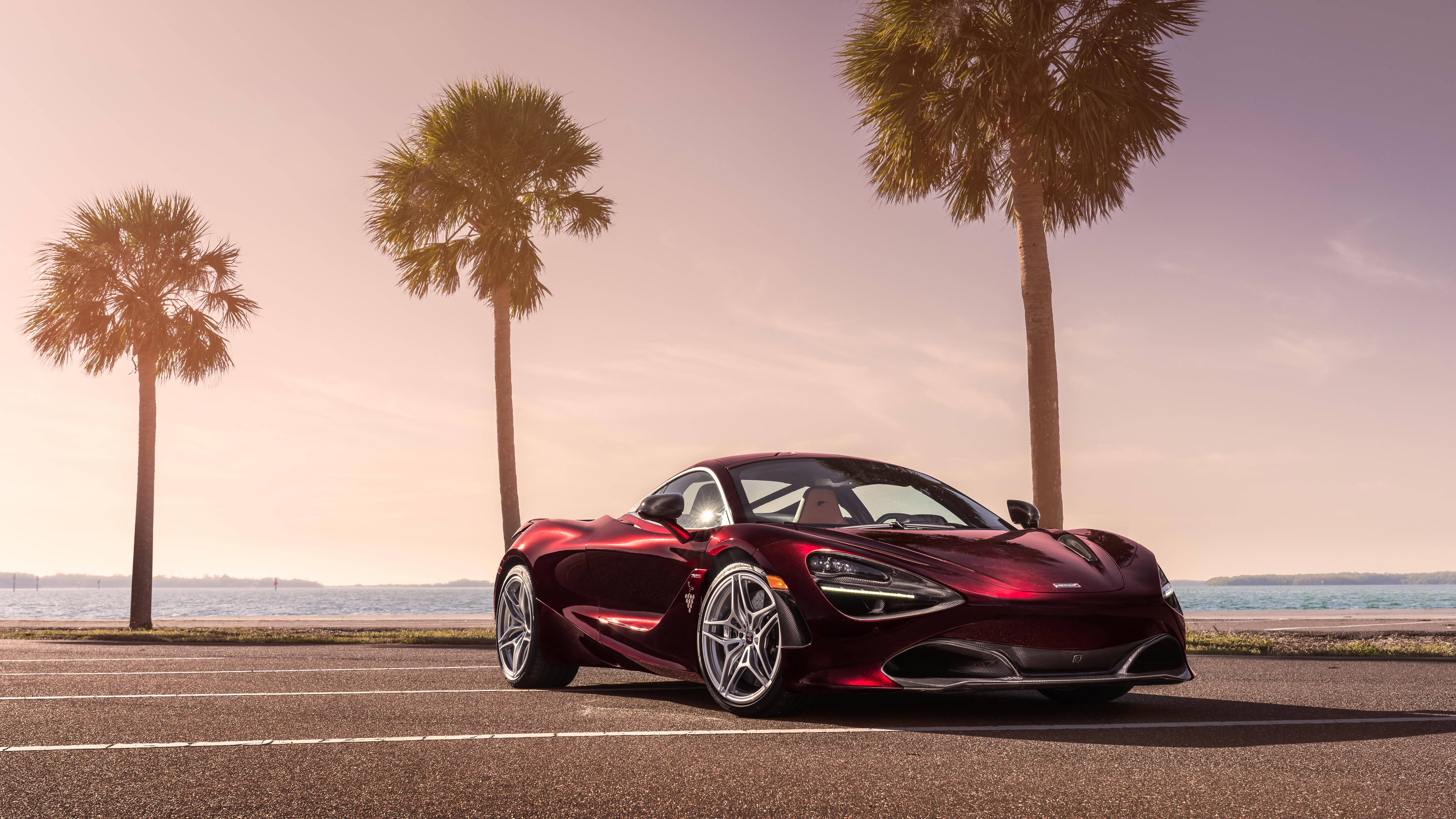 Mclaren 720s 桌布 壁紙 — 一輛亮紅色McLaren 720S跑車停泊在日落時分的海岸道路上，背景有棕櫚樹和海洋。