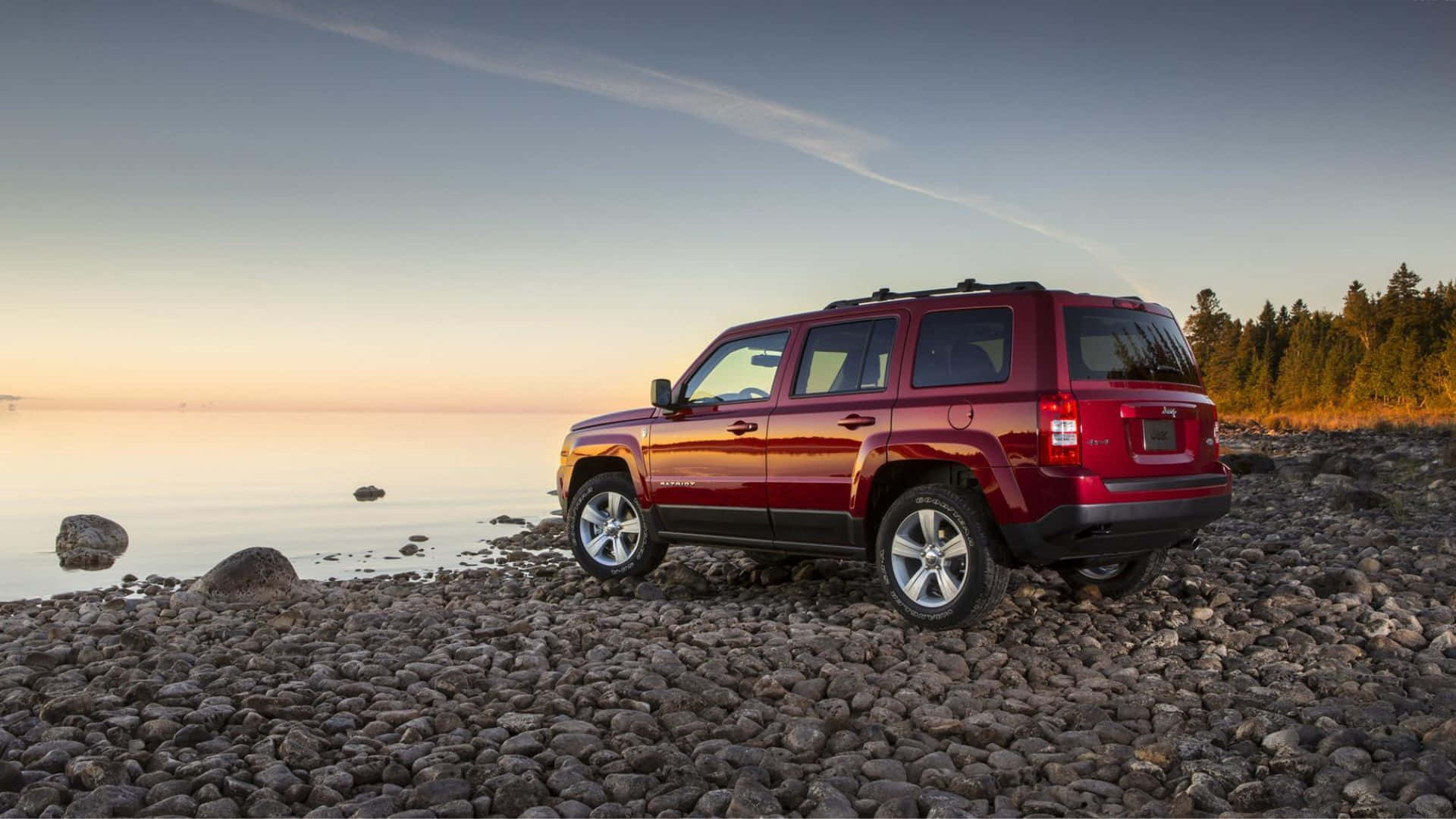 Jeep Patriot 桌布 壁紙 — 一輛亮紅色SUV停泊在日落時分的岩石湖岸邊，平靜的水面和遠處的樹木在柔和漸變的天空下。