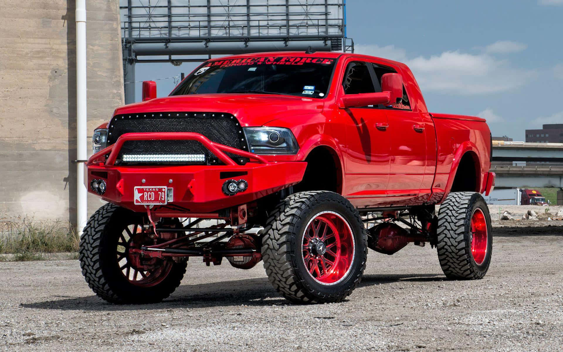 Jacked Up Trucks 桌布 壁紙 — 一輛亮紅色升高底盤的皮卡車，配備超大輪胎和定制格柵，停在工業建築附近的礫石上，天空局部多雲。