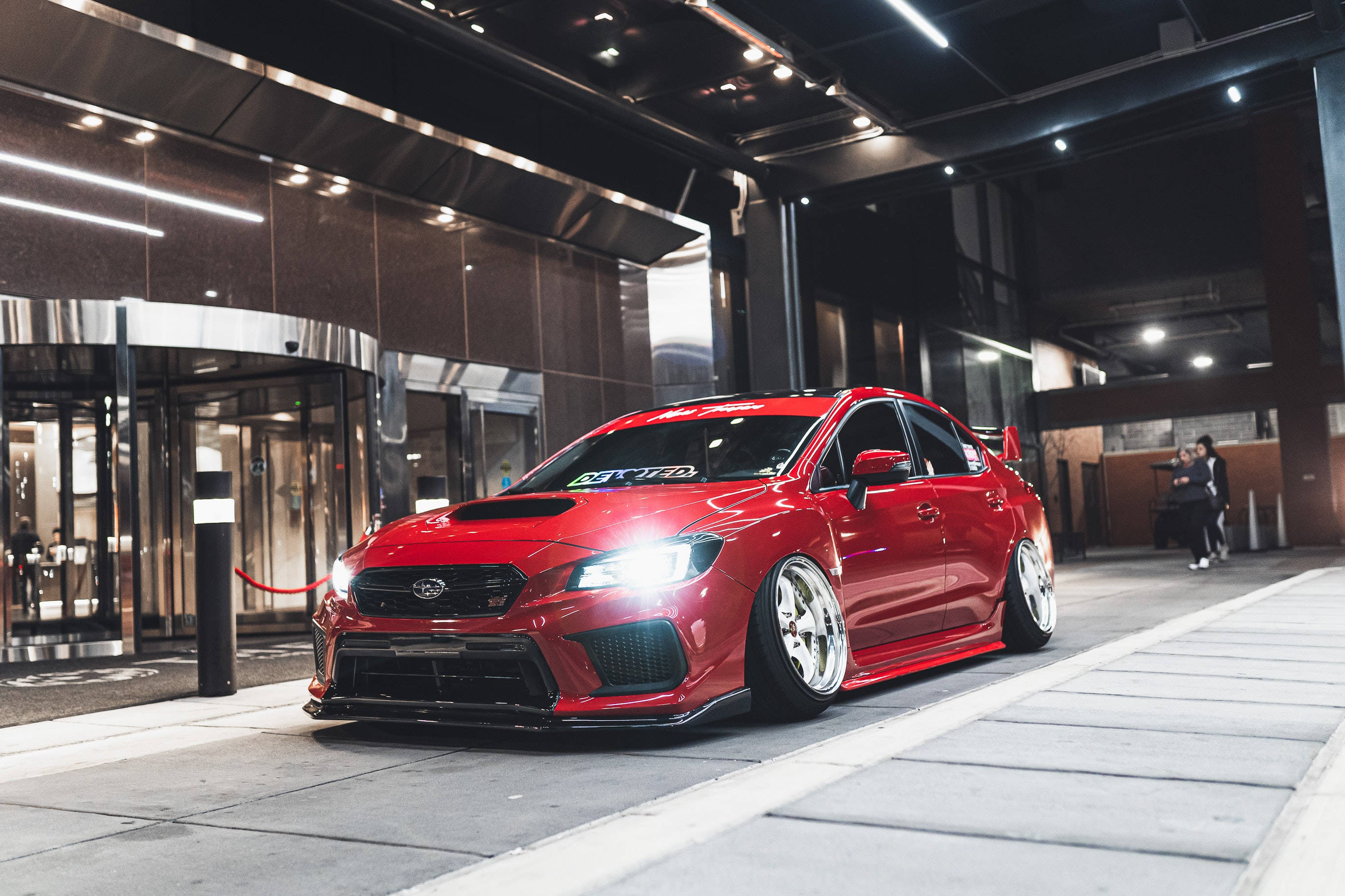 昴 桌布 壁紙 — 一輛亮紅色改裝Subaru WRX STI夜晚停在現代建築物外面，車頭燈亮著。