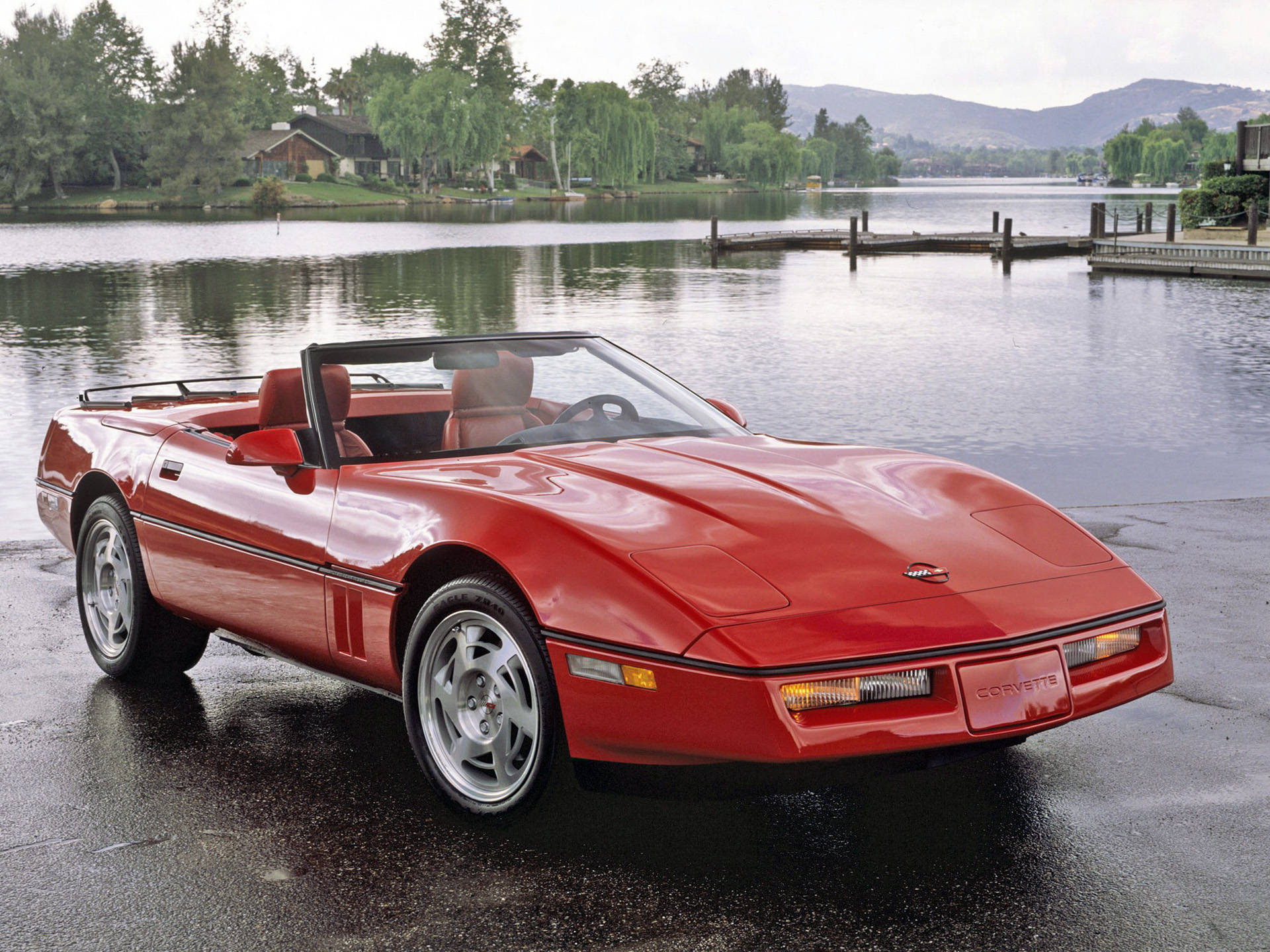corvette 桌布 壁紙 — 一輛亮紅色敞篷跑車停泊在濕滑的碼頭上，旁邊是一個平靜的湖泊，背景有樹木和山丘。