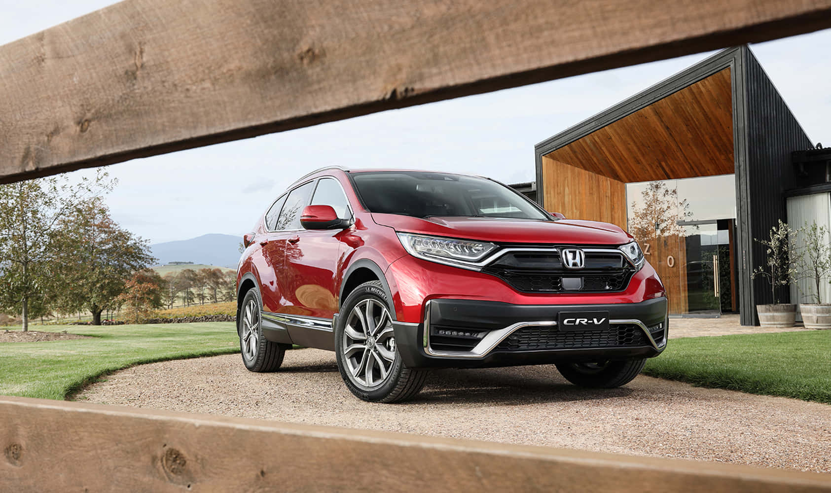 Honda Cr-v 桌布 壁紙 — 一輛亮紅色本田CR-V SUV停放在碎石車道上，車道由木樑框起，背景是一座現代房屋和連綿起伏的山丘。