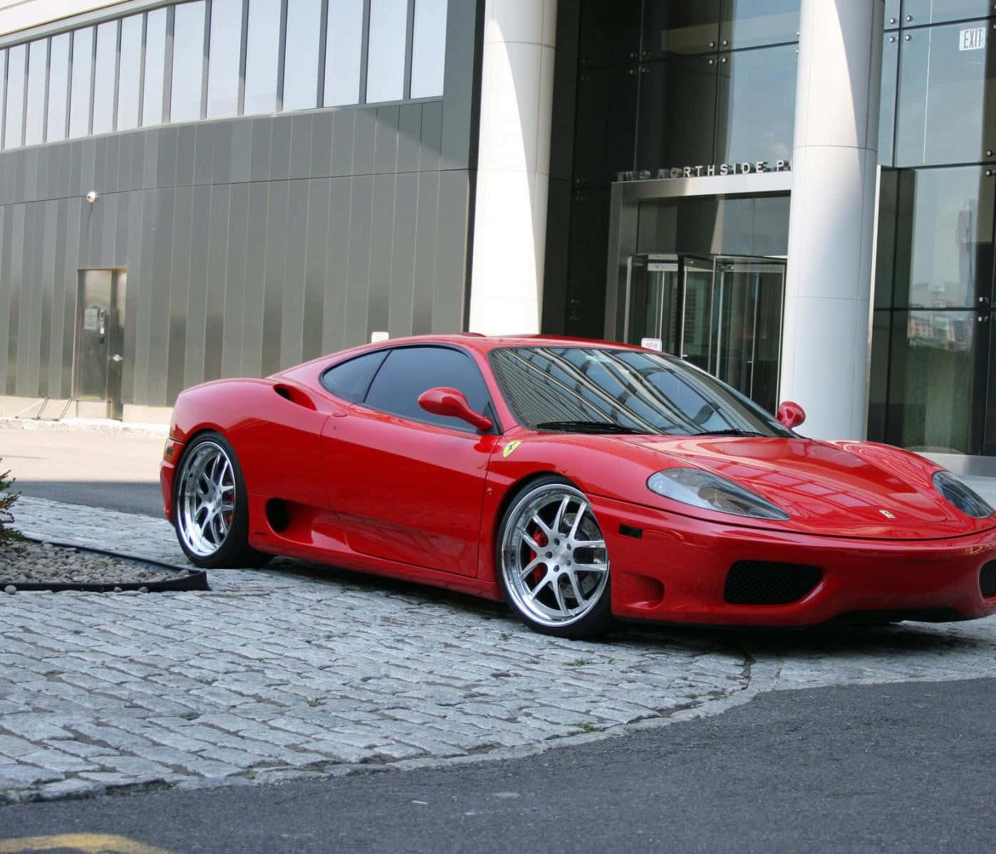 Ferrari 360 Modena 桌布 壁紙 — 一輛亮紅色法拉利360跑車停放在現代玻璃建築前的鵝卵石車道上。