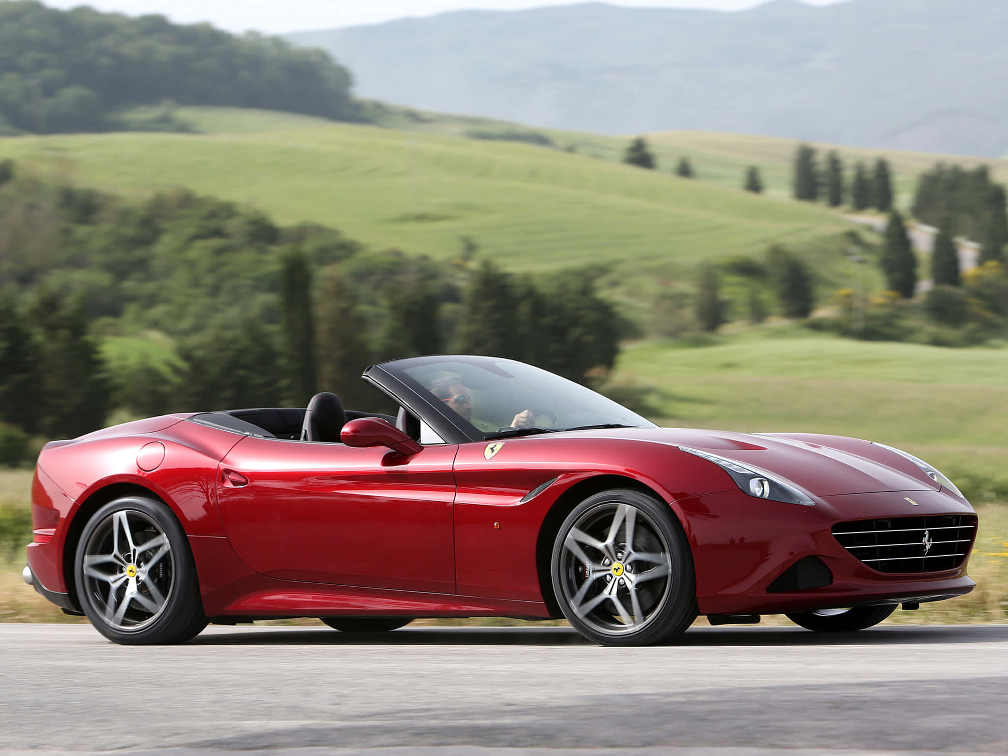 Ferrari California T 桌布 壁紙 — 一輛亮紅色法拉利California敞篷車在明亮的日光下沿著蜿蜒的道路穿過鬱鬱蔥蔥的綠色山丘行駛。