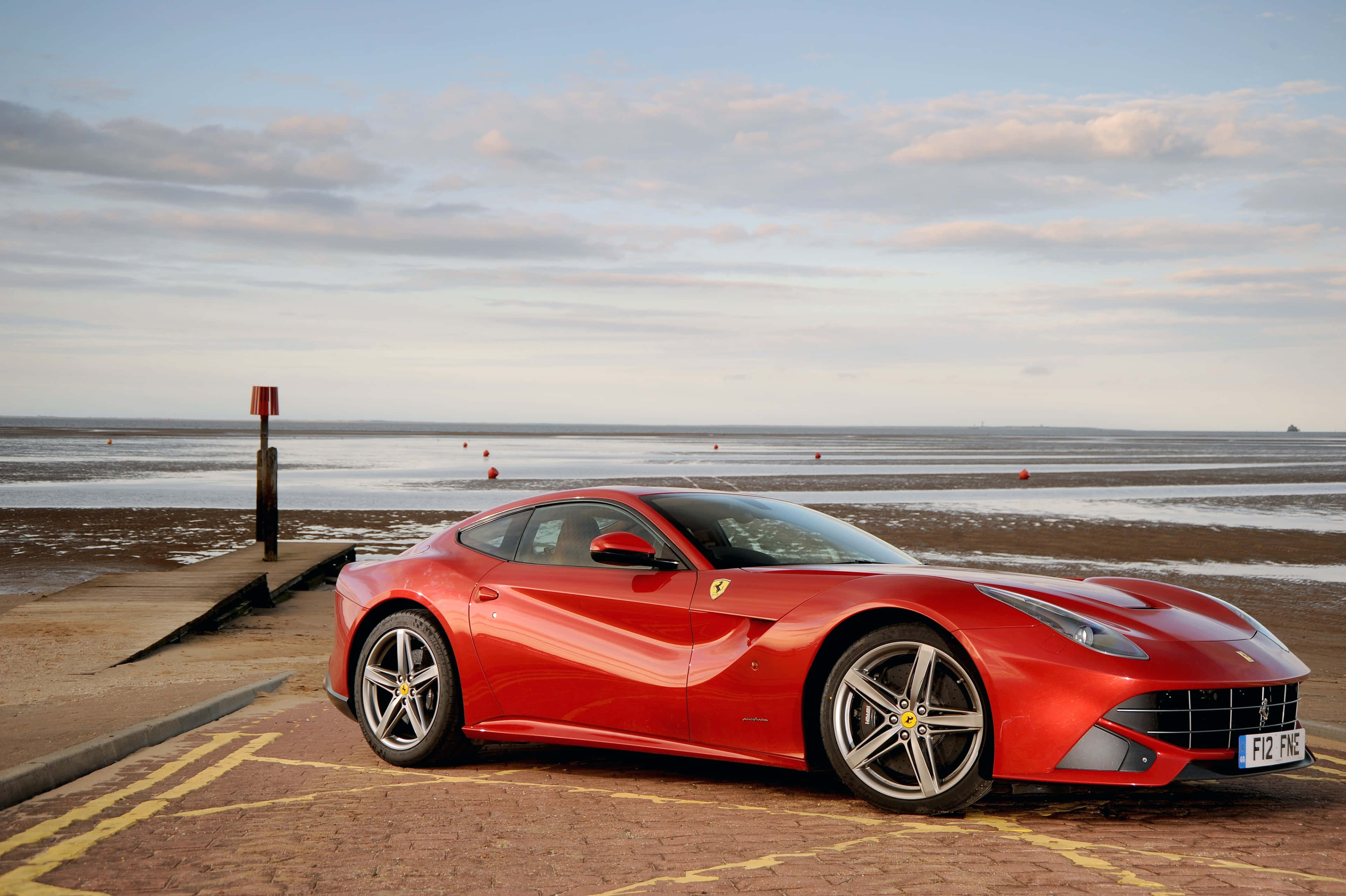 Ferrari F12 Berlinetta 桌布 壁紙 — 一輛亮紅色法拉利F12停泊在磚砌碼頭上，背景是日落時分的平靜海面和散落的浮標。