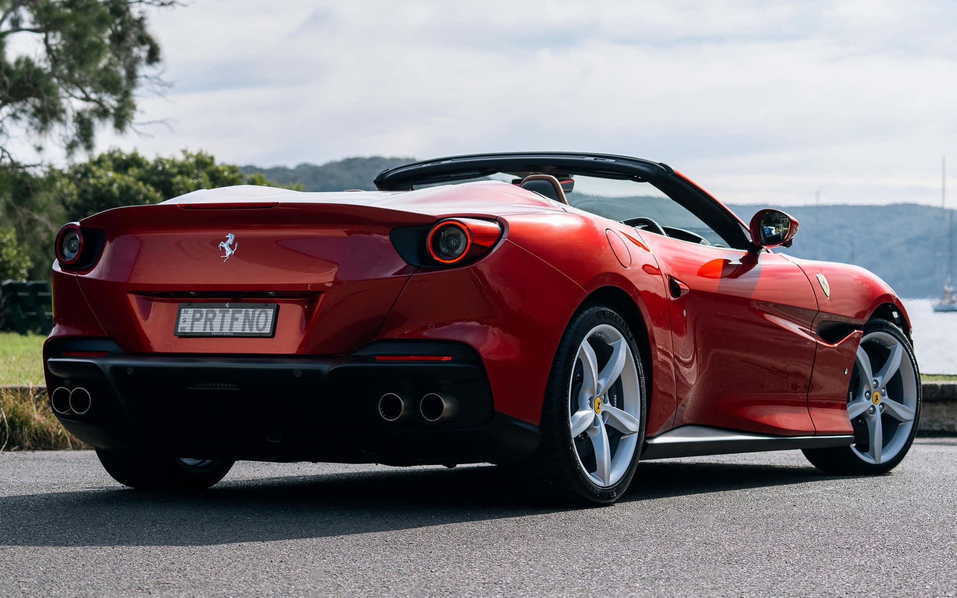 Ferrari Portofino 桌布 壁紙 — 一輛亮紅色法拉利Portofino敞篷跑車停泊在沿海道路上，背景有樹木和水面。