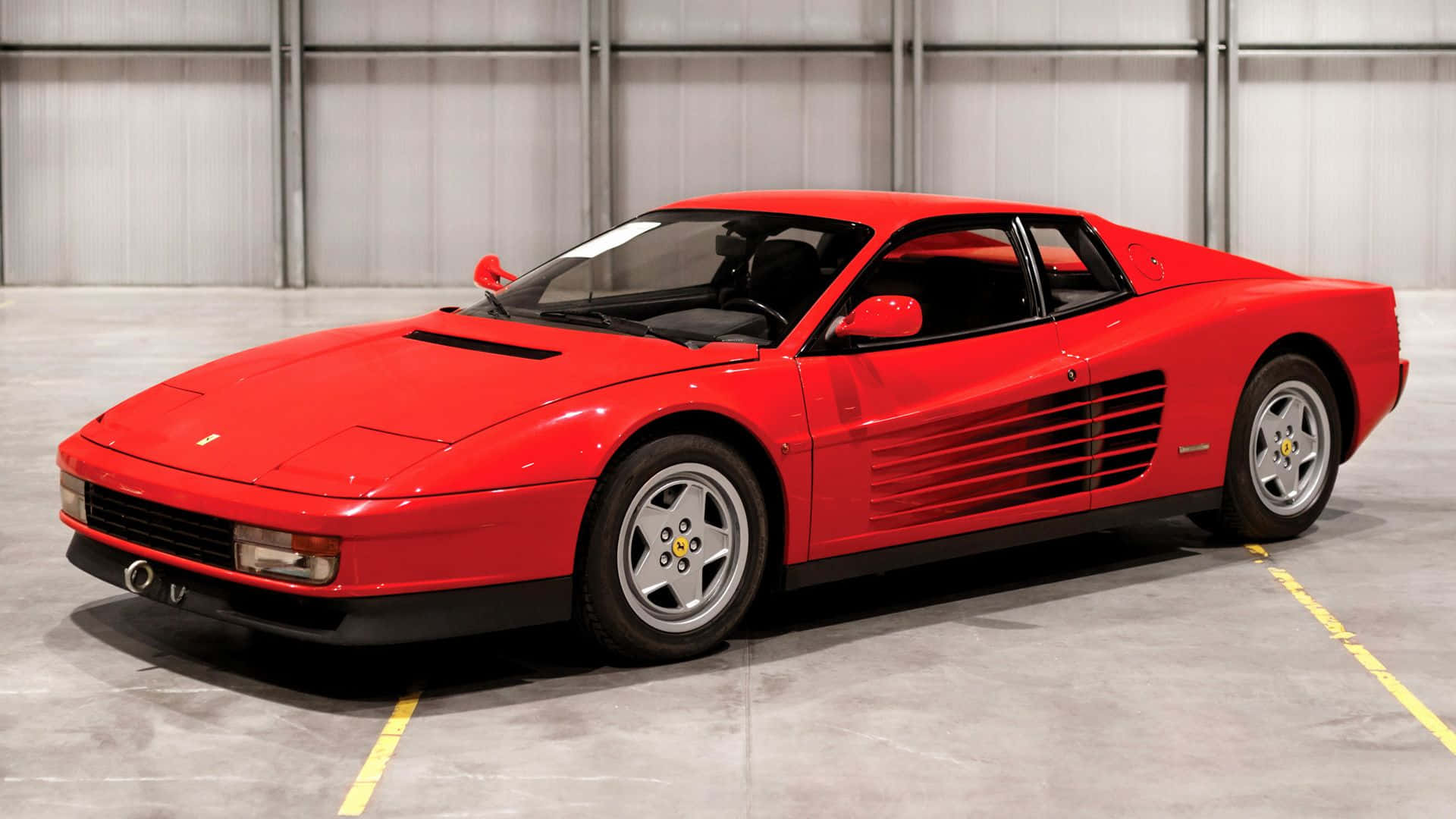 Ferrari Testarossa 桌布 壁紙 — 一輛亮紅色法拉利Testarossa停放在寬敞的工業車庫中，車庫有混凝土地板和金屬牆壁。