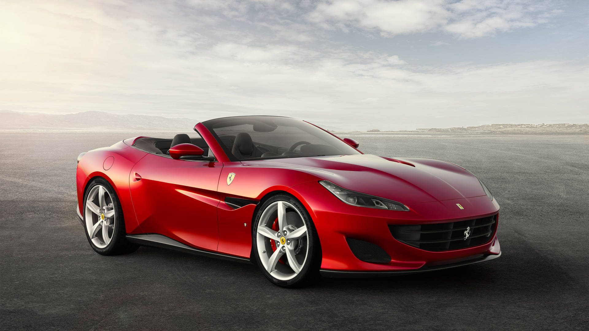 1920x1080 Ferrari 桌布 壁紙 — 一輛亮紅色法拉利敞篷跑車停放在廣闊的灰色瀝青場地上，天空柔和多雲。