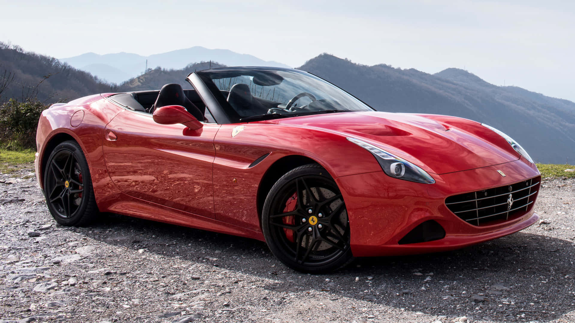 Ferrari California T 桌布 壁紙 — 一輛亮紅色法拉利敞篷車停在碎石路上，背景是起伏的丘陵和遠山，天空晴朗。