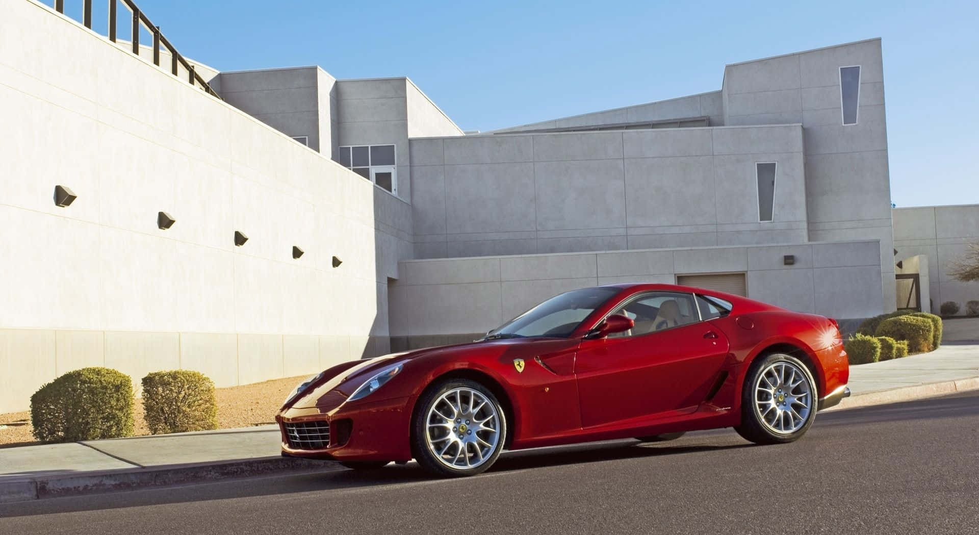 Ferrari 599 Gtb Fiorano 桌布 壁紙 — 一輛亮紅色法拉利跑車停放在街道上，旁邊是現代混凝土建築，天空清澈湛藍。
