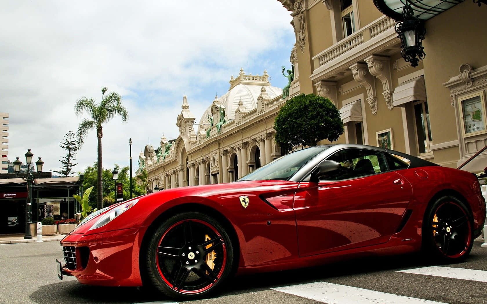 Ferrari 599 Gtb Fiorano 桌布 壁紙 — 一輛亮紅色法拉利跑車停泊在街上，旁邊是一棟有棕櫚樹的裝飾性米色建築，天空局部多雲。