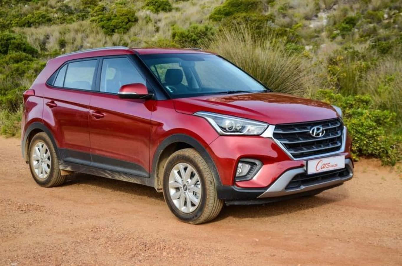 Hyundai 桌布 壁紙 — 一輛亮紅色現代SUV停在泥路上，周圍是綠色灌木和乾草，在明亮的日光下。