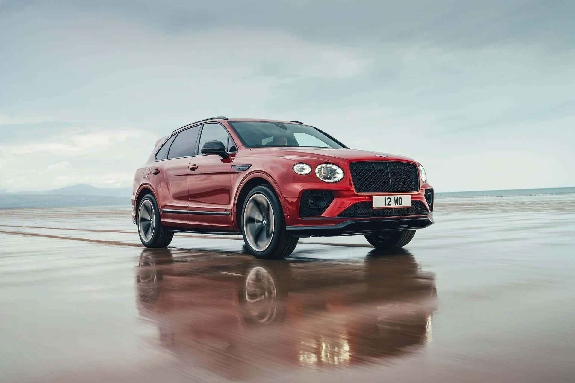 Bentley Bentayga 桌布 壁紙 — 一輛亮紅色的 Bentley Bentayga SUV 停在濕漉漉的海灘上，在多雲的天空下反射出流線型的外形。