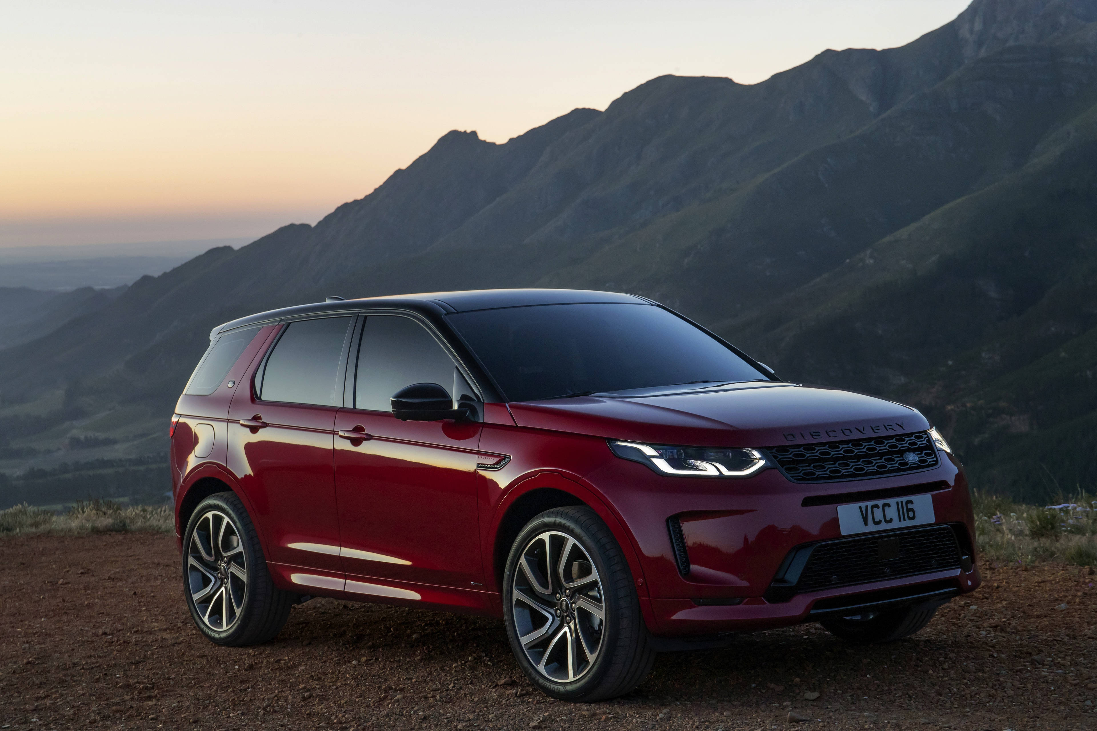 land rover iPhone 桌布 壁紙 — 一輛亮紅色的 Land Rover Discovery 停在一個 dirt 觀景點，背景是起伏的綠色山脈和柔和的日落天空。