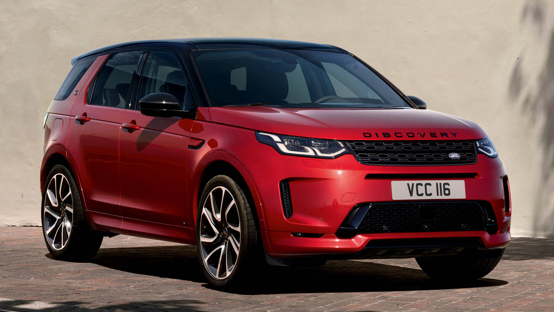 land rover iPhone 桌布 壁紙 — 一輛亮紅色的 Land Rover Discovery SUV 停泊在磚砌路面上，背景是純色米色牆壁，在明亮日光下。