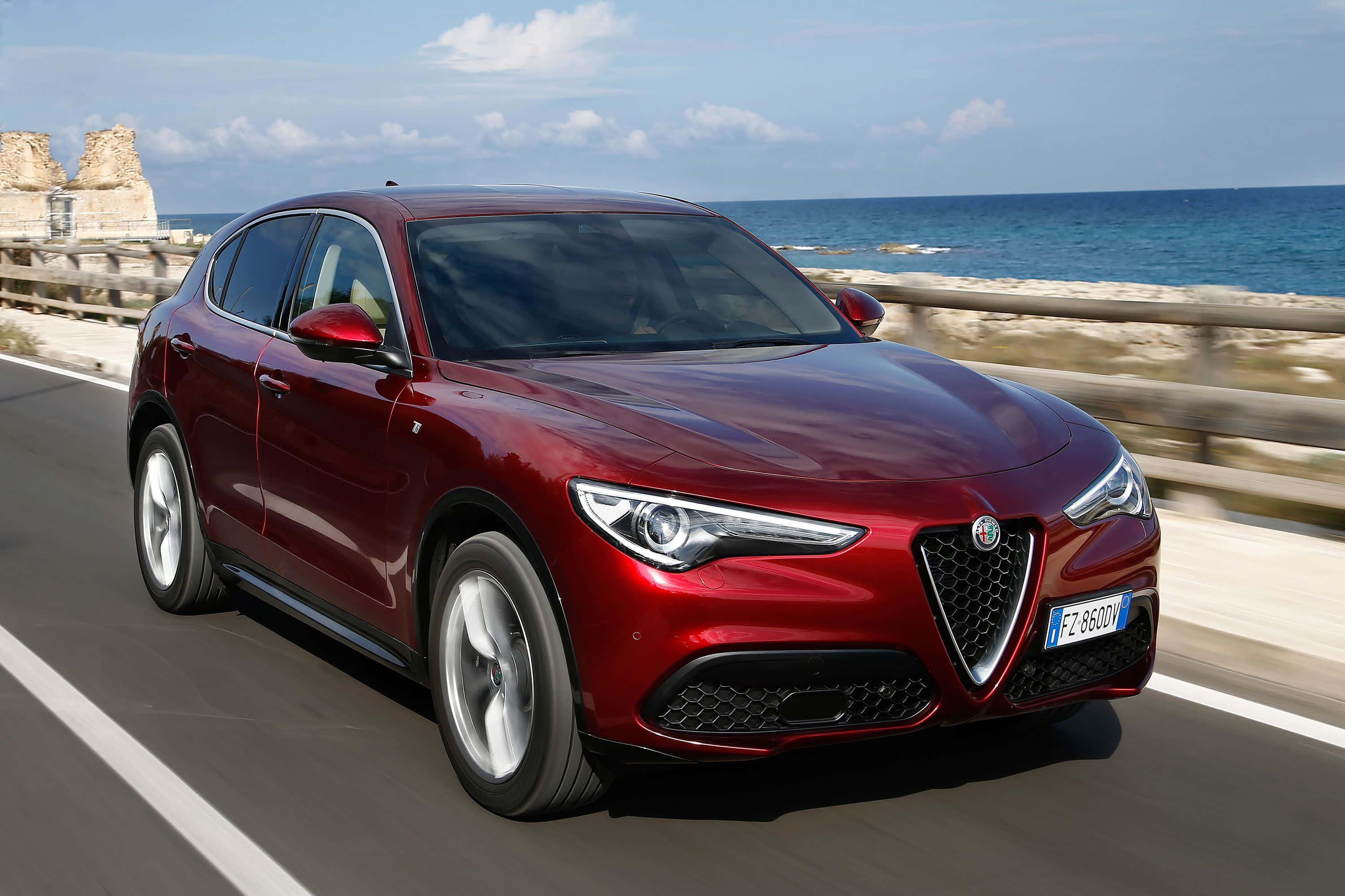 愛快羅密歐 桌布 壁紙 — 一輛亮紅色的Alfa Romeo SUV沿著海岸公路飛馳，背景是藍色的海洋和天空。