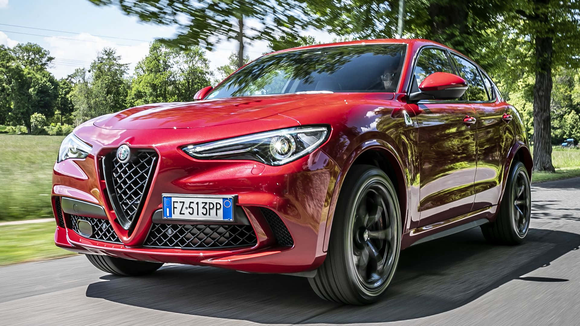 Alfa Romeo Stelvio 桌布 壁紙 — 一輛亮紅色的Alfa Romeo SUV在夏日陽光下沿著綠樹成蔭的道路疾馳。