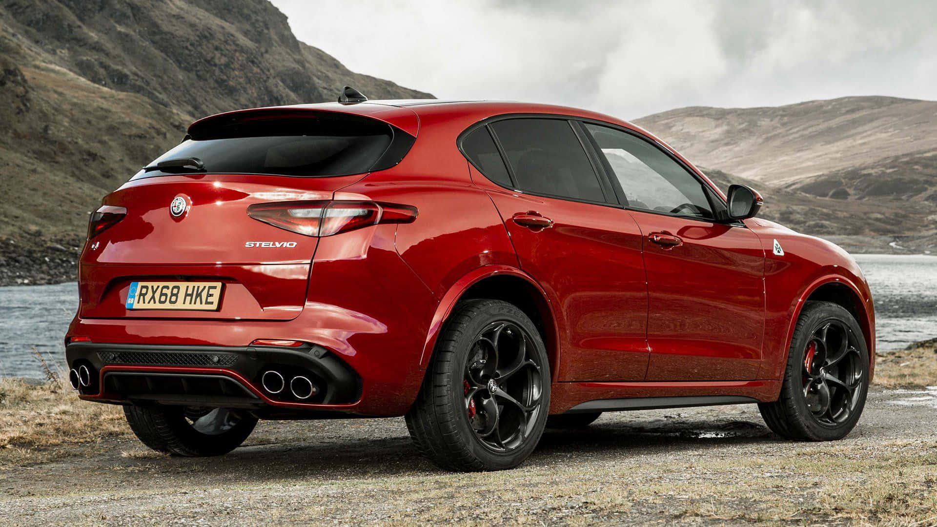Alfa Romeo Stelvio 桌布 壁紙 — 一輛亮紅色的Alfa Romeo Stelvio SUV停泊在碎石岸邊，旁邊是平靜的湖泊，背景有崎嶇的山丘，天空多雲。