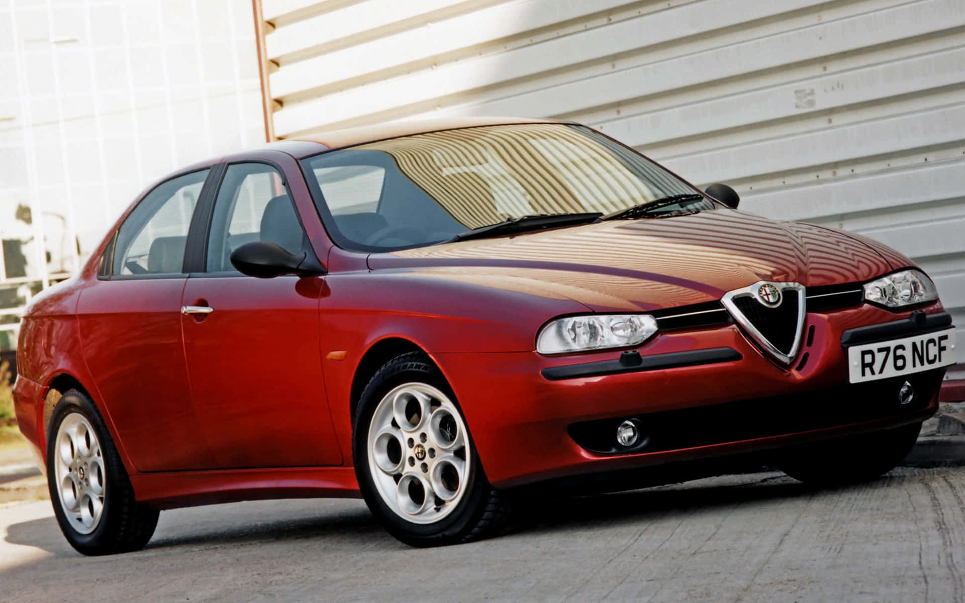 Alfa Romeo 156 桌布 壁紙 — 一輛亮紅色的Alfa Romeo 156轎車在明亮的日光下以傾斜角度停放在白色波紋金屬牆旁。