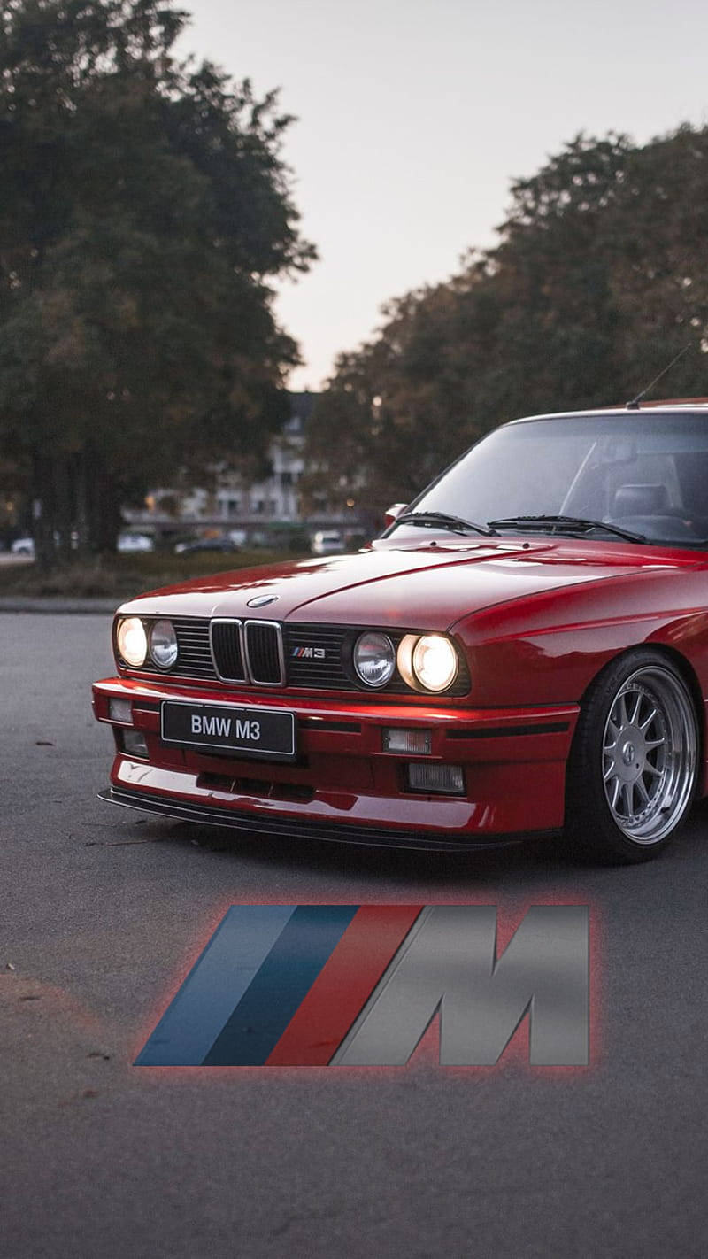 m 桌布 壁紙 — 一輛亮紅色的BMW M3跑車停放在黃昏時分的林蔭大道上，車頭燈亮起，M標誌在人行道上發光。