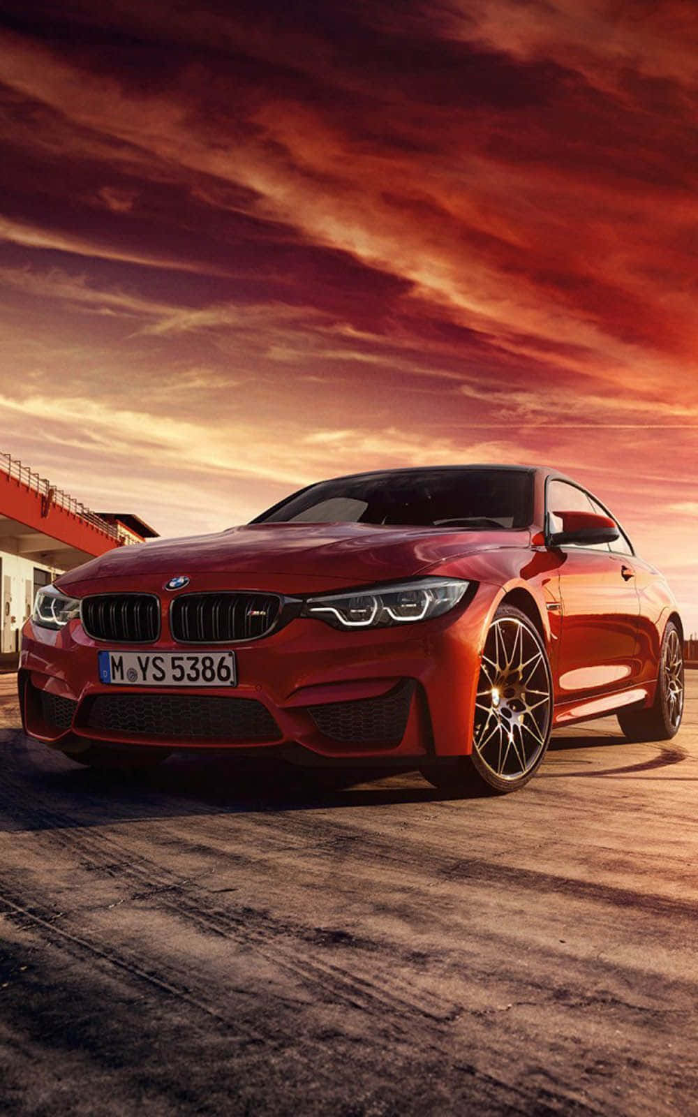 BMW 動圖 桌布 壁紙 — 一輛亮紅色的BMW M4雙門跑車停放在混凝土停車場上，背景是火熱的橙紫色日落天空。