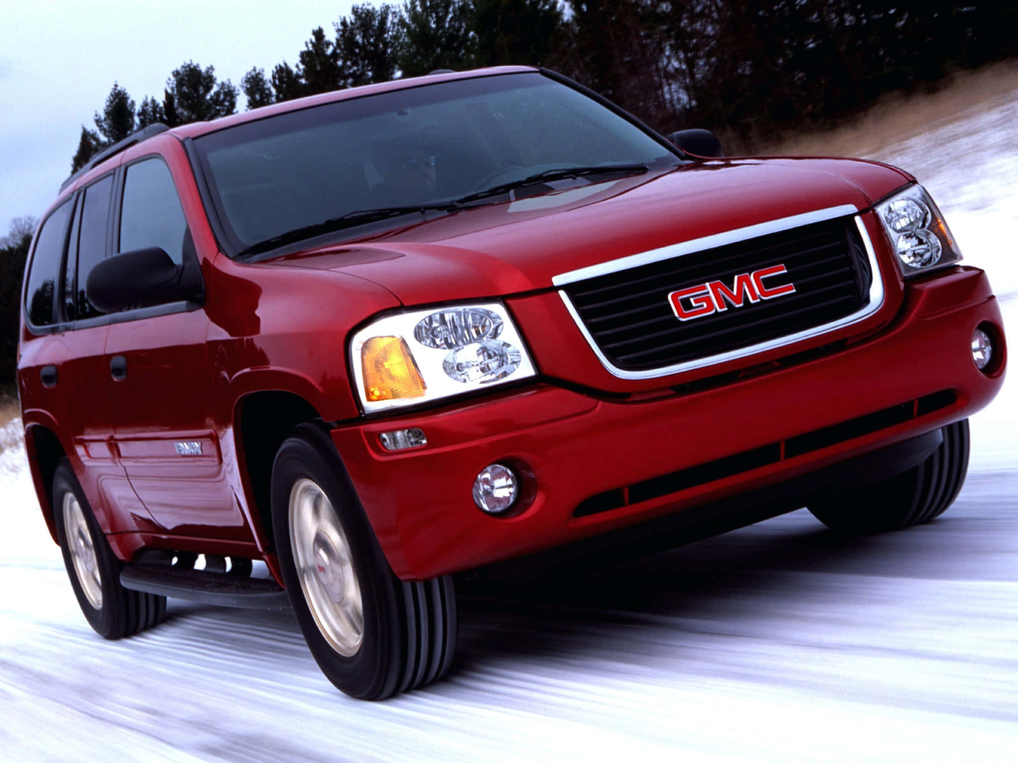 Gmc Envoy 桌布 壁紙 — 一輛亮紅色的GMC Envoy SUV在雪地中行駛，車輪帶有動態模糊效果。
