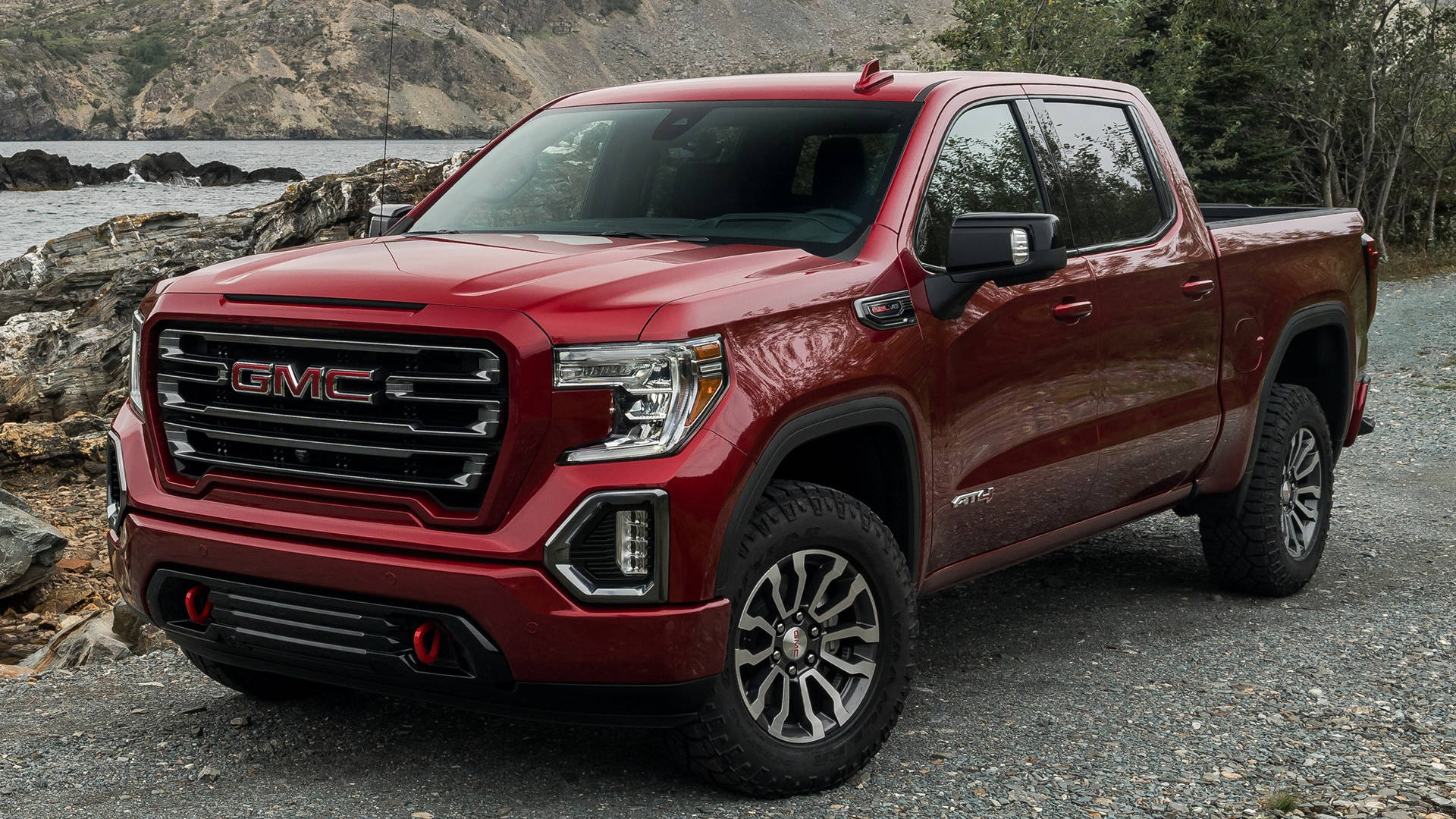 Gmc 桌布 壁紙 — 一輛亮紅色的GMC Sierra AT4皮卡車停在碎石上，靠近岩石海岸線，背景有水和樹木。